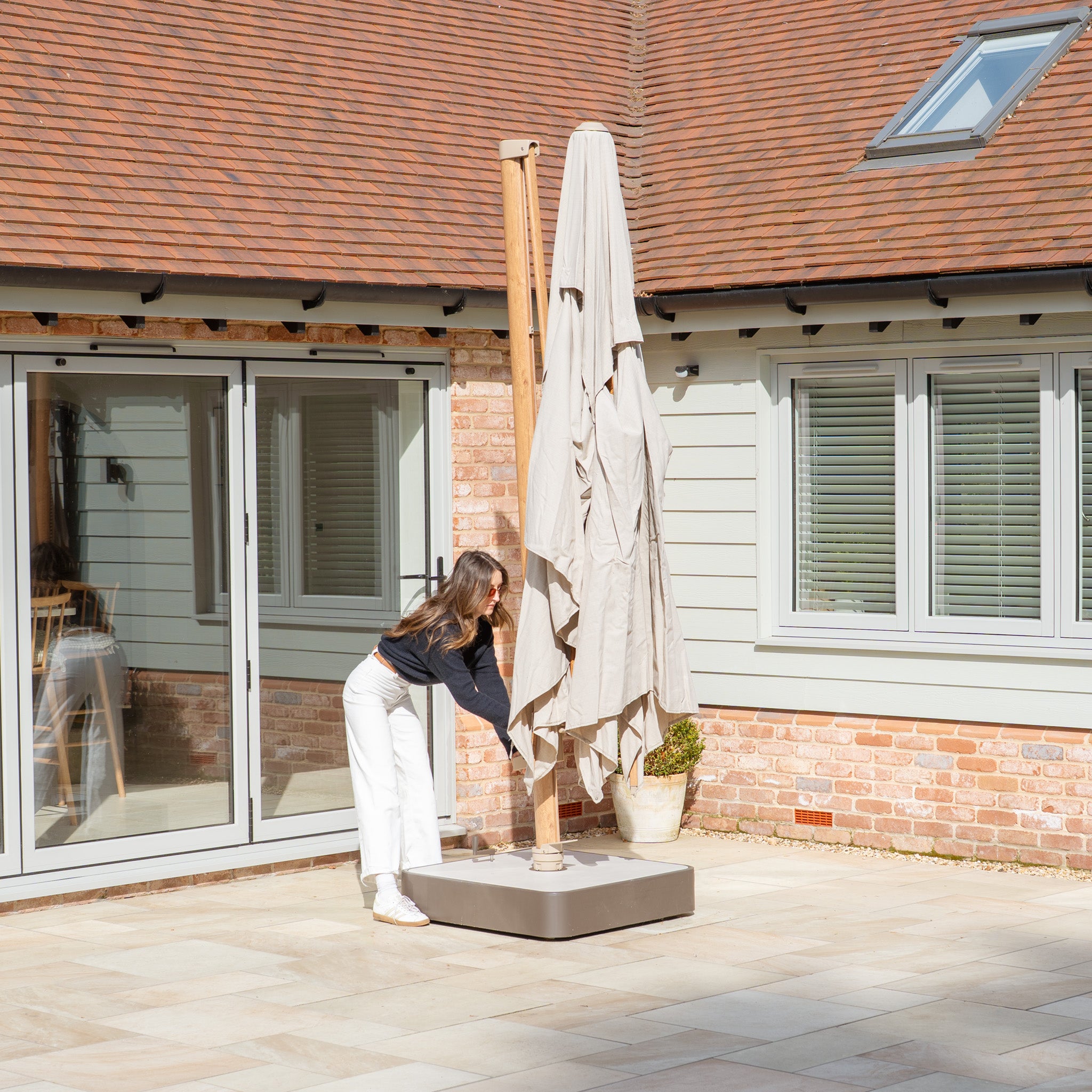On the patio of a cozy brick and wood house, someone adjusts the Finca 4m x 3m Cantilever Parasol in Sand for optimal sun protection, enhancing outdoor living.