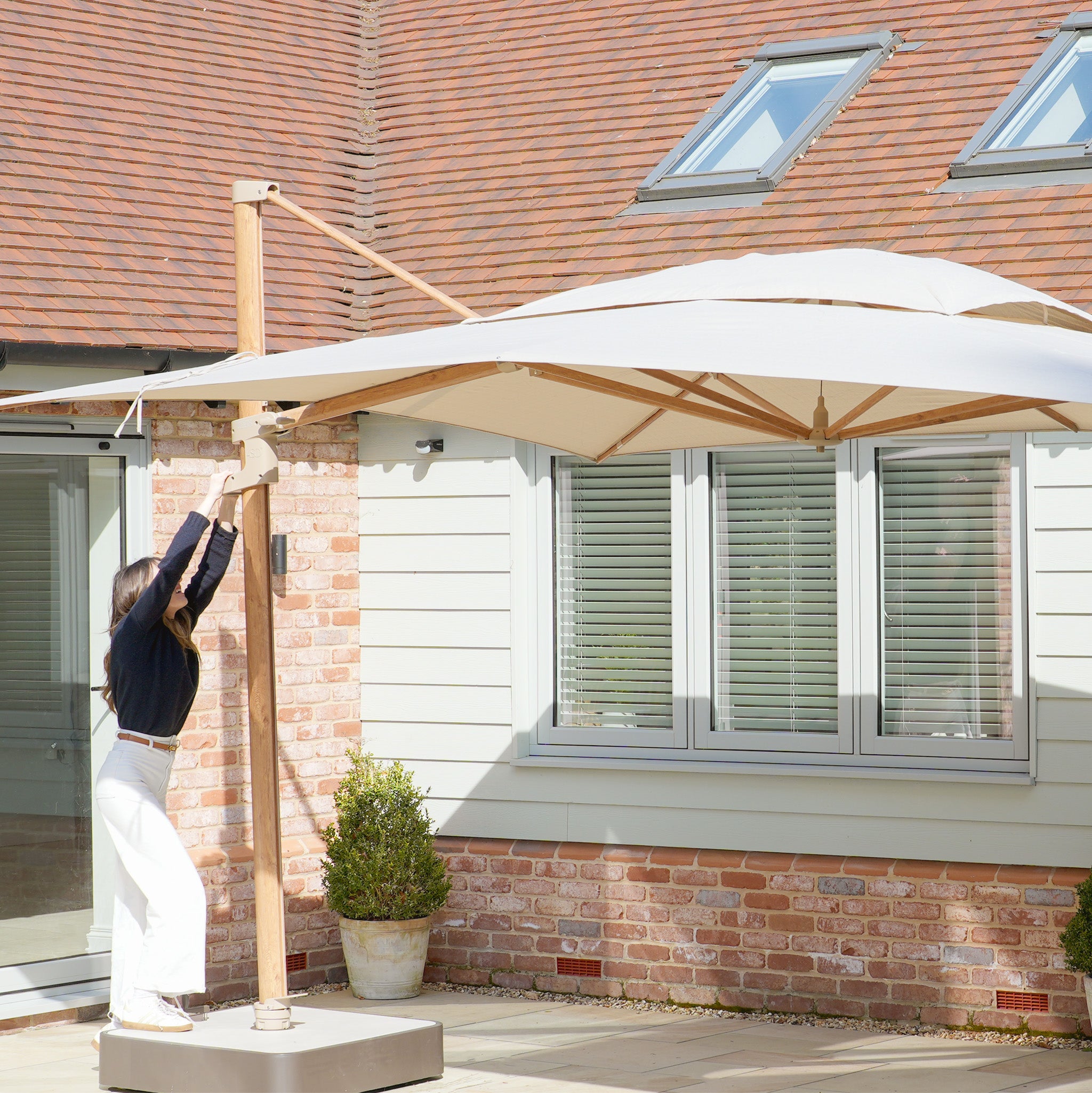 In a modern brick and siding courtyard, someone expertly adjusts the Finca 4m x 3m Cantilever Parasol in Sand, boosting outdoor living among lush potted plants and ensuring great sun protection.