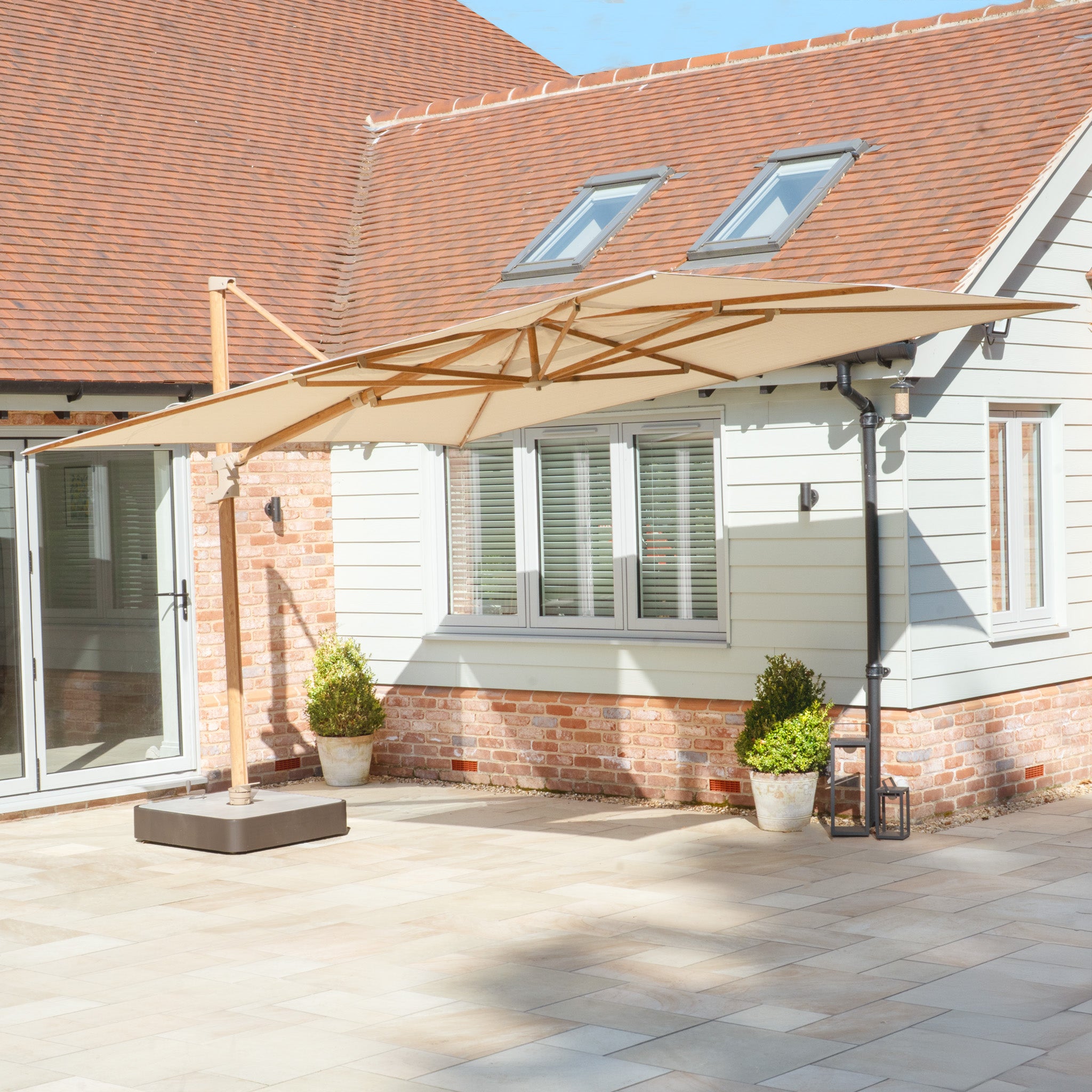 Patio area with a Finca 4m x 3m Cantilever Parasol in Sand made from durable olefin fabric, providing sun protection. It's surrounded by potted plants and set against a brick and siding house exterior.