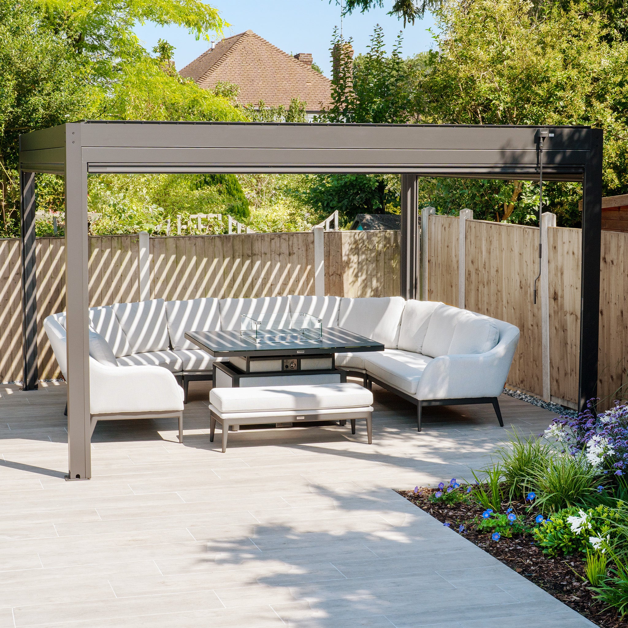The inviting outdoor seating area features white sofas under the sleek PergoSTET Plus 4m x 4m Square Pergola in Grey with subtle LED lighting and three drop sides, all set on a tiled patio surrounded by lush trees and vibrant flowers.
