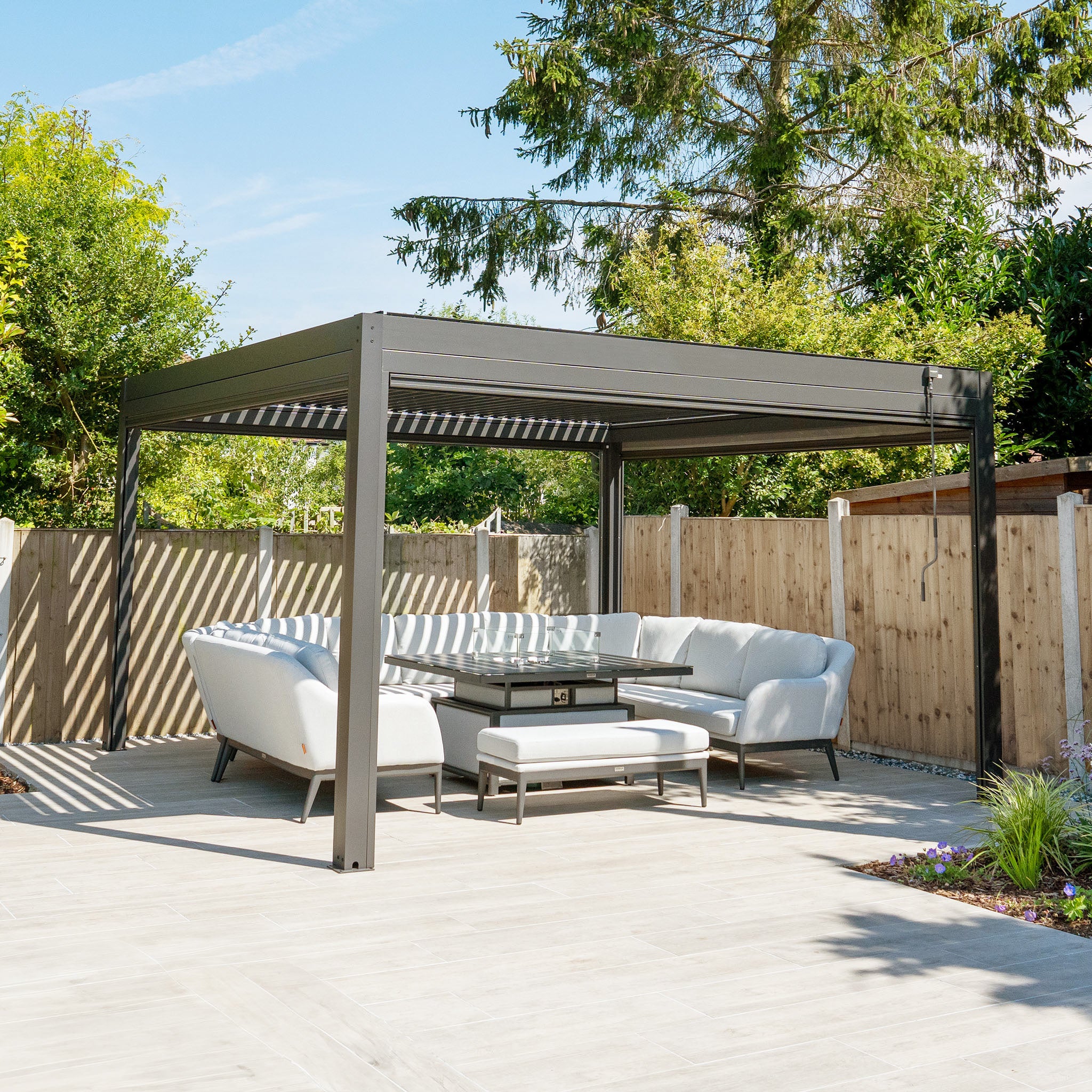 A modern patio featuring the PergoSTET Plus 4m x 4m Square Pergola with 3 Drop Sides and LED Lighting in Grey is enhanced by a white sectional sofa, creating an ideal outdoor living space with a sleek table, wooden fence, and lush greenery.