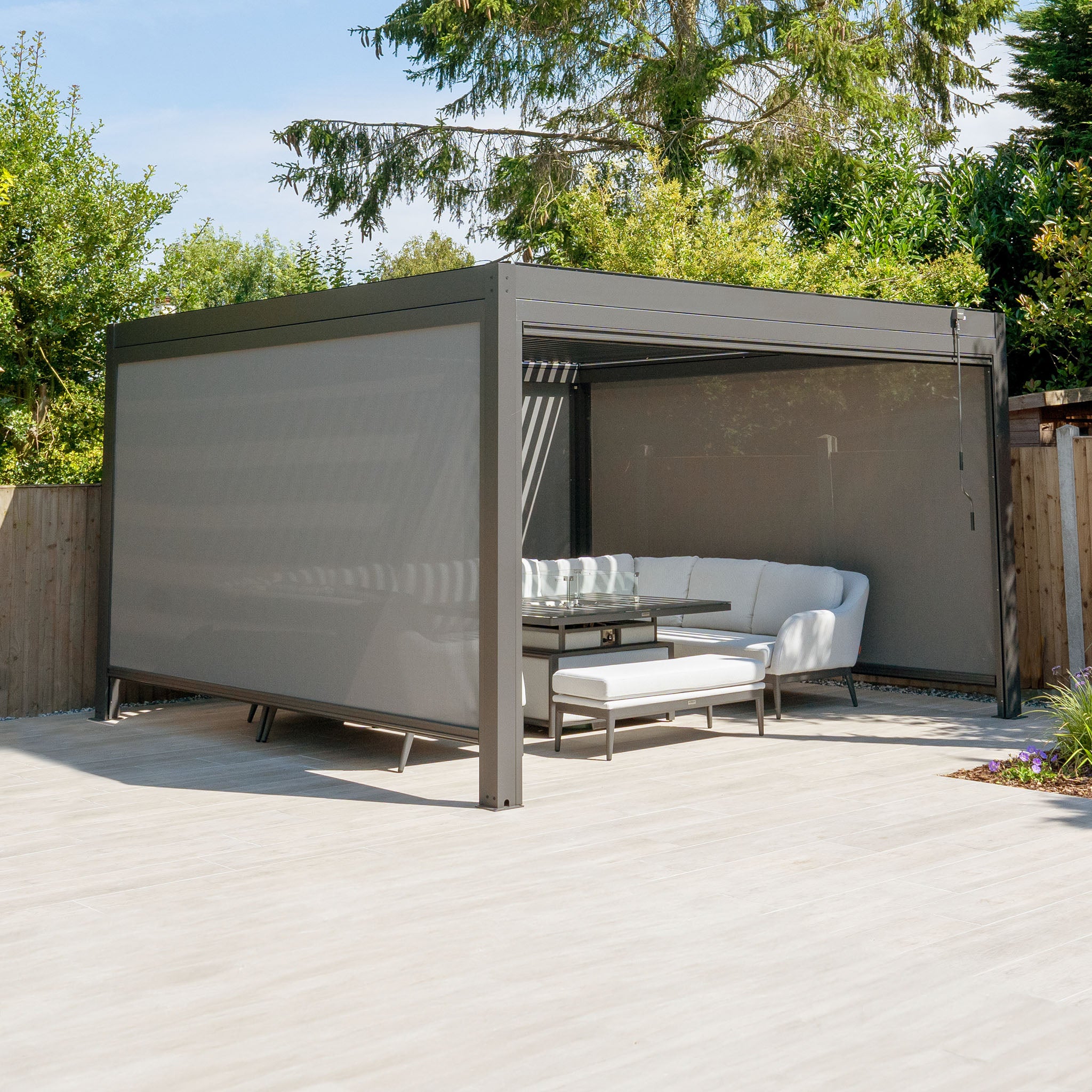 An inviting outdoor living space features the PergoSTET Plus 4m x 4m Square Pergola in Grey with LED lighting and three drop sides on a wooden deck. A white sectional sofa and glass table add modern comfort, while surrounding trees enhance its appeal.