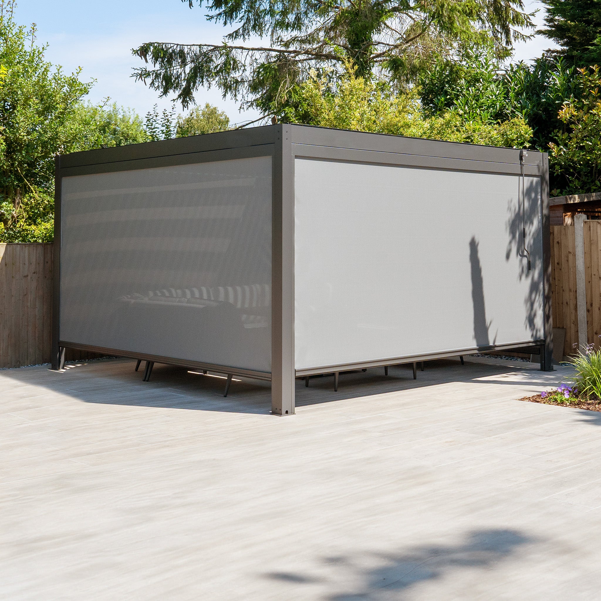 The PergoSTET Plus 4m x 4m Square Pergola, featuring three drop sides and LED lighting in grey, enhances the sunlit patio's outdoor living space amid trees and a wooden fence with its modern design.