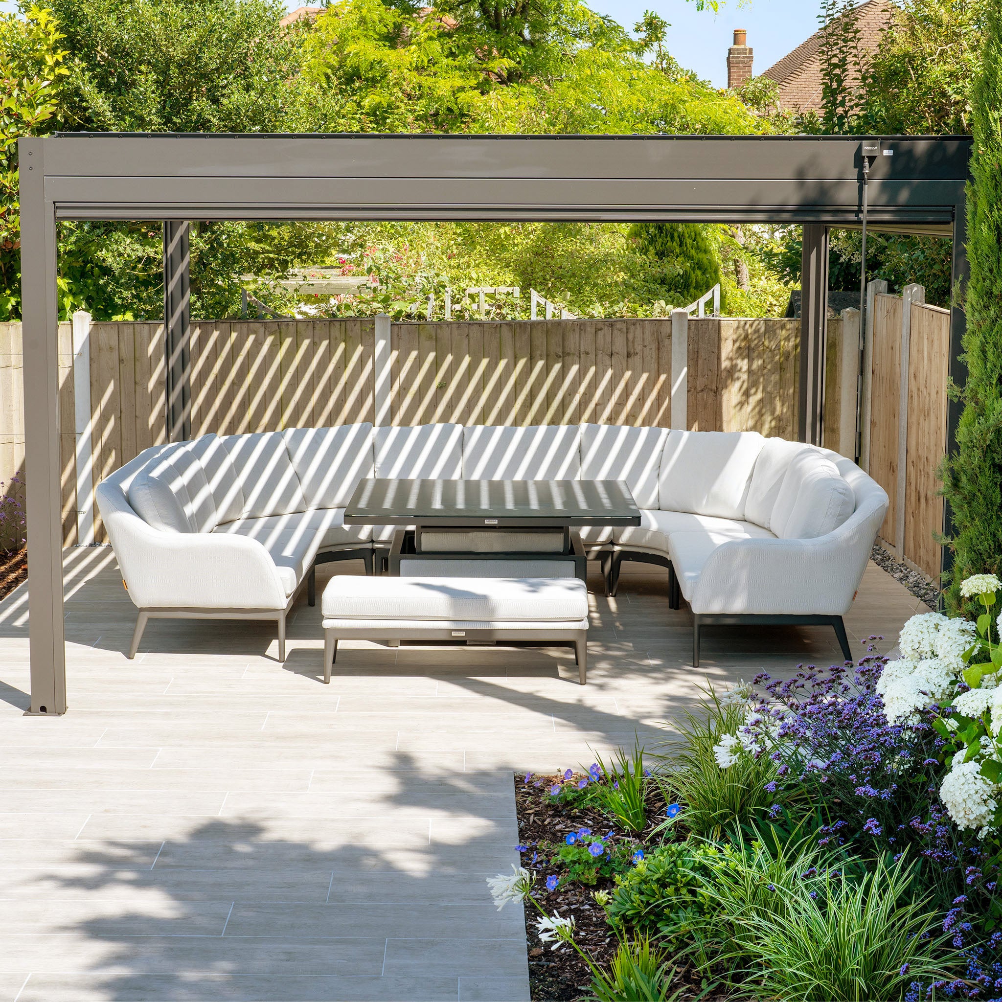 A modern outdoor patio features the Luna U-Shape Outdoor Fabric Corner Dining Set with a Rising Table in Oyster Grey and a pergola, all set in a sunlit garden.