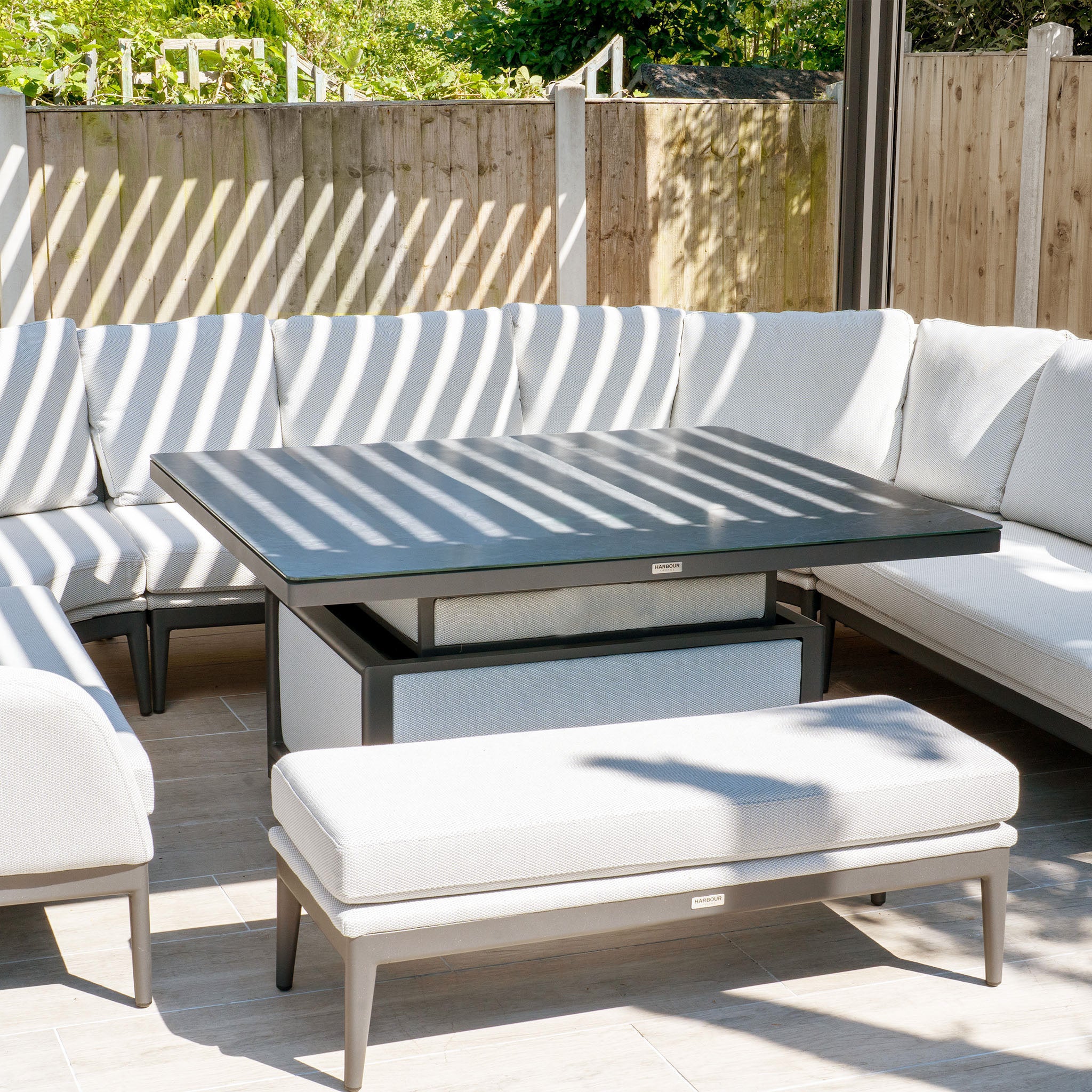 The Luna U-Shape Outdoor Fabric Corner Dining features an aluminium frame and beige cushions, paired with a dark rising firepit table. Wooden fencing enhances the setting, creating an ideal space for relaxation and conversation.