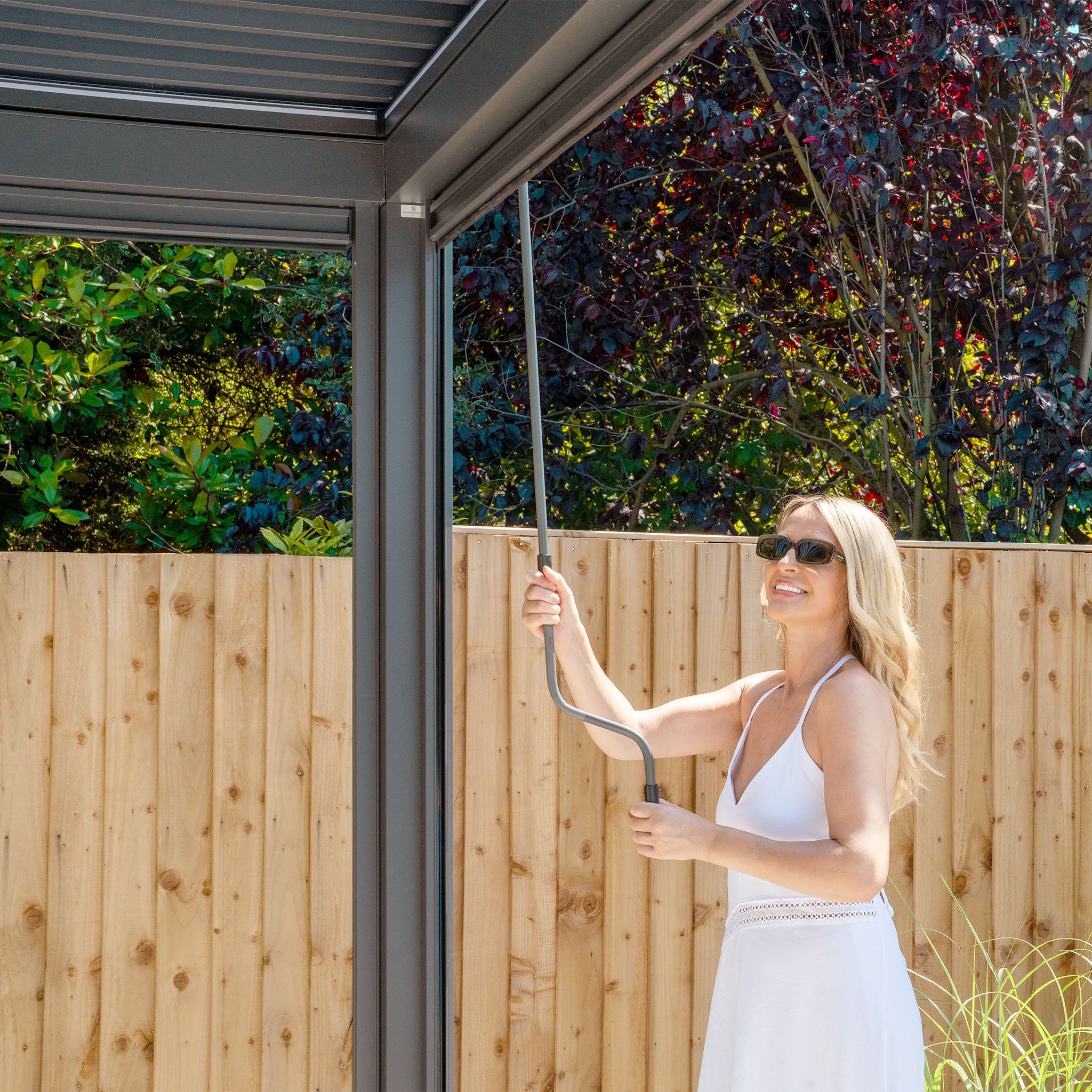 A woman in a white dress uses a metal rod to adjust the PergoSTET Plus 4m x 4m Square Pergola with 3 drop sides and LED lighting in Grey, enhancing her outdoor living space against a wooden fence and lush foliage backdrop.