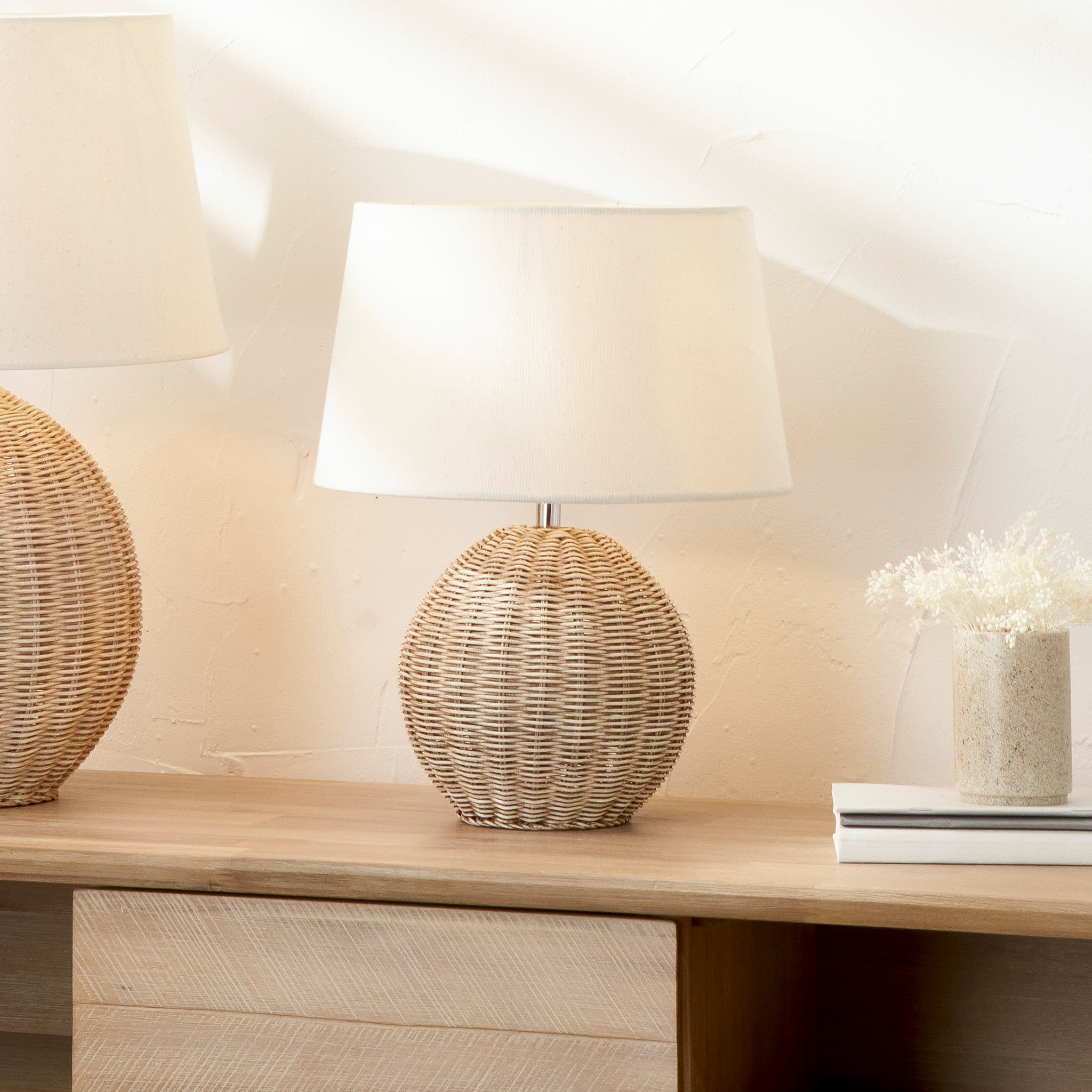 The Pacific Lifestyle - Raffles Small Rattan Cream Wash Table Lamp, featuring an antique cream wash rattan base and cream slubby cotton shade, sits on a wooden surface beside a vase of dried flowers and books, evoking the charm of a cozy beach hut retreat.