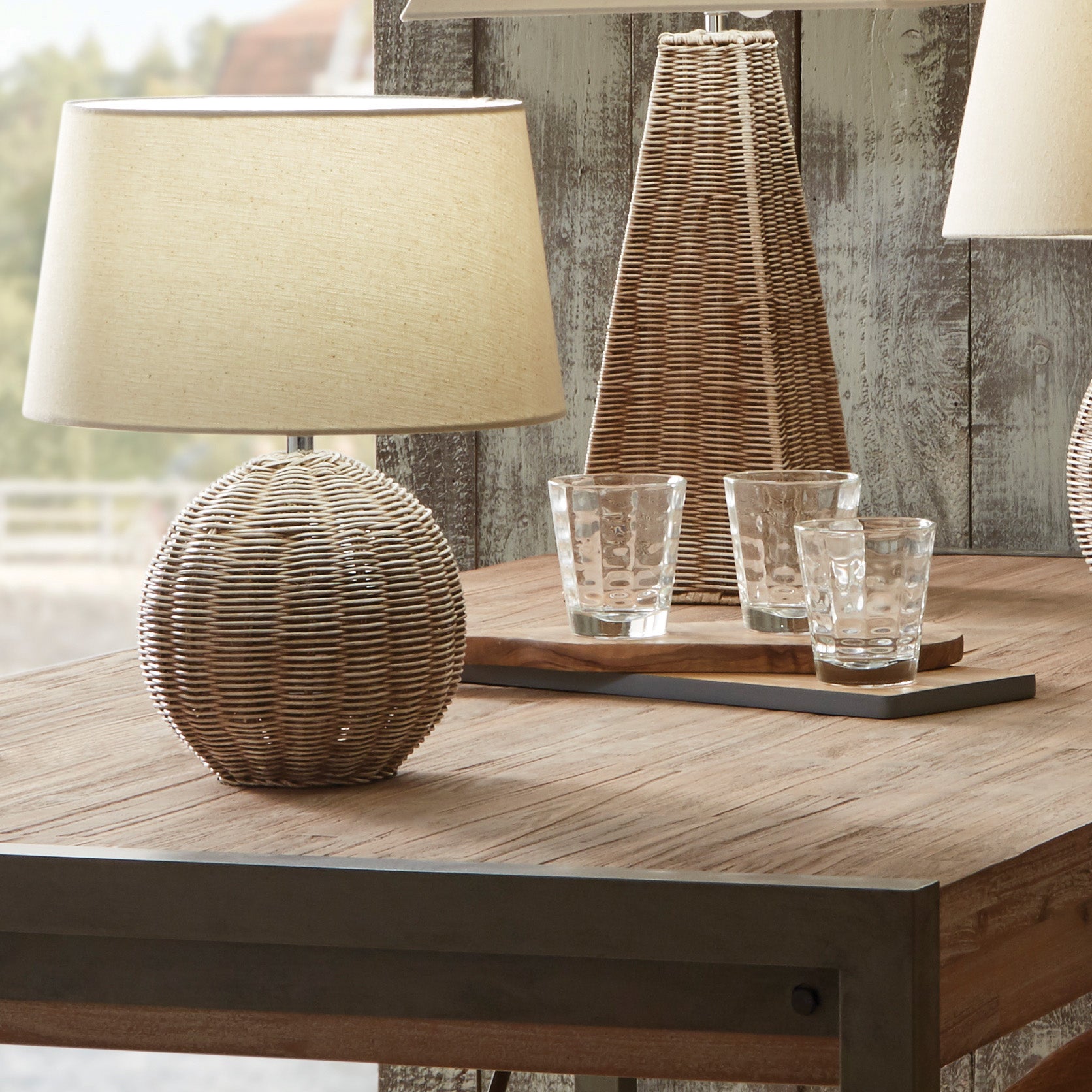 The Pacific Lifestyle Raffles Small Rattan Cream Wash Table Lamp, combined with a cream slubby cotton shade and two glasses on a wooden table, offers a rustic decor vibe reminiscent of a cozy beach hut.