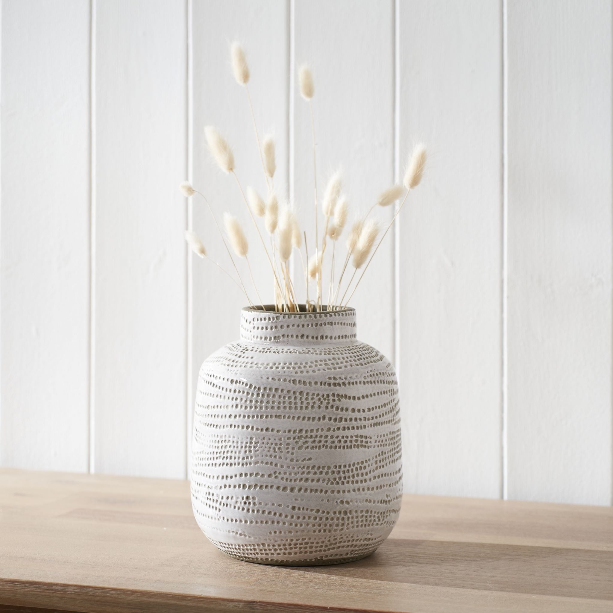 The Pacific Lifestyle Alina Dot Design Small Stoneware Vase in White, filled with dried grasses, sits elegantly on a wooden table against a white paneled wall.