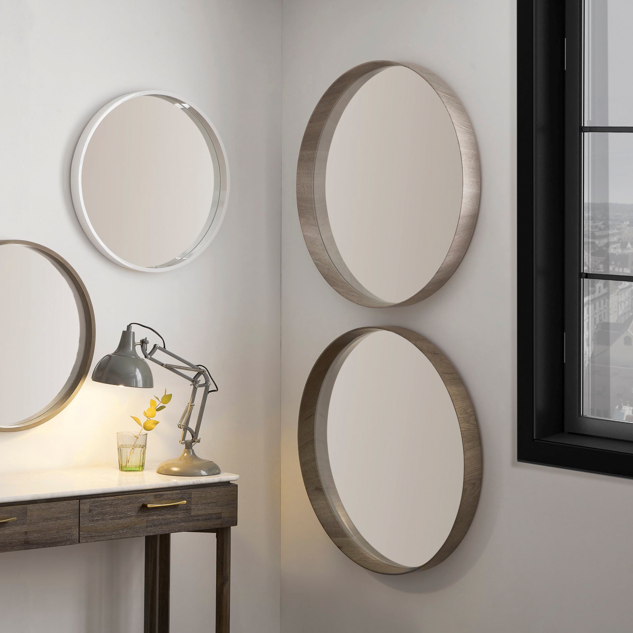 A minimalist modern corner boasts a Pacific Lifestyle Natural Wood Veneer Round Wall Mirror, a natural wood desk, sleek metal lamp, and glass for your drink. The window reveals a vibrant city view adding urban flair to the serene space.