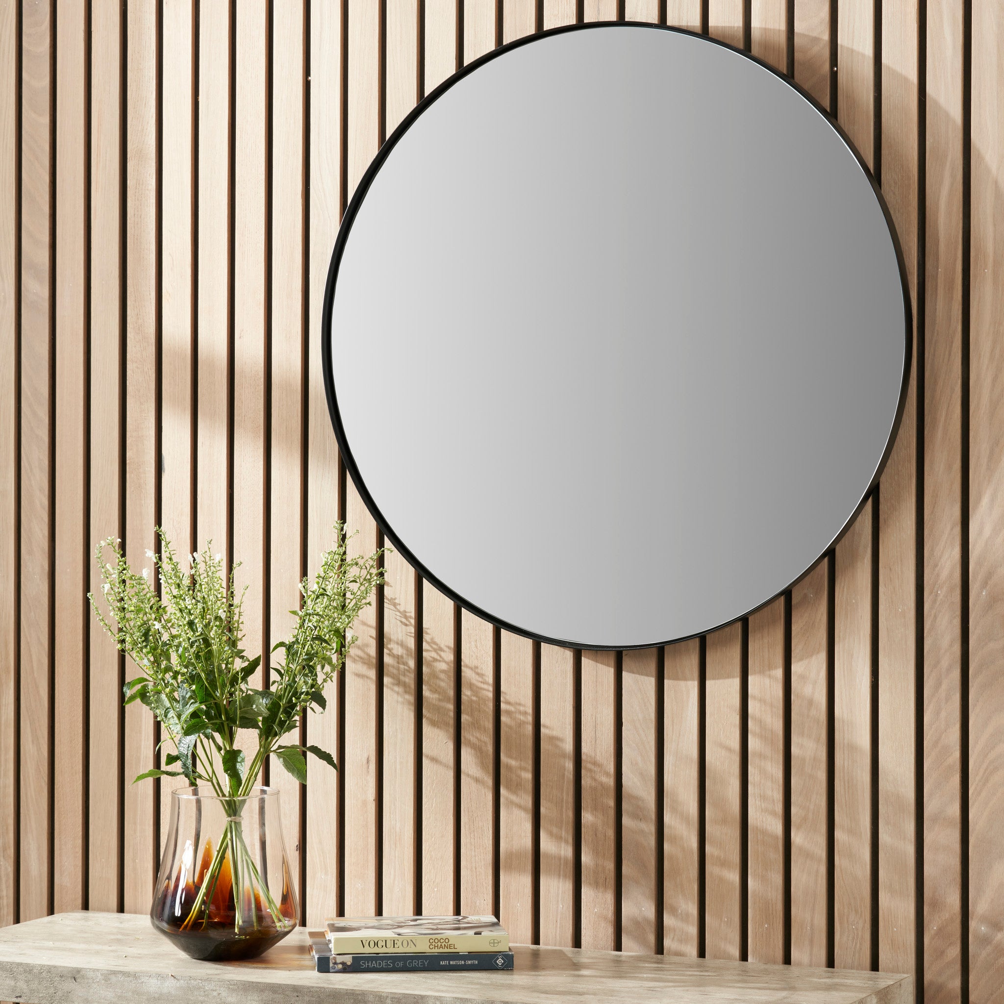 A Pacific Lifestyle Black Metal Slim Frame Round Wall Mirror adorns the wood-paneled wall, complemented by a vase of flowers and two books elegantly positioned on the concrete table below.