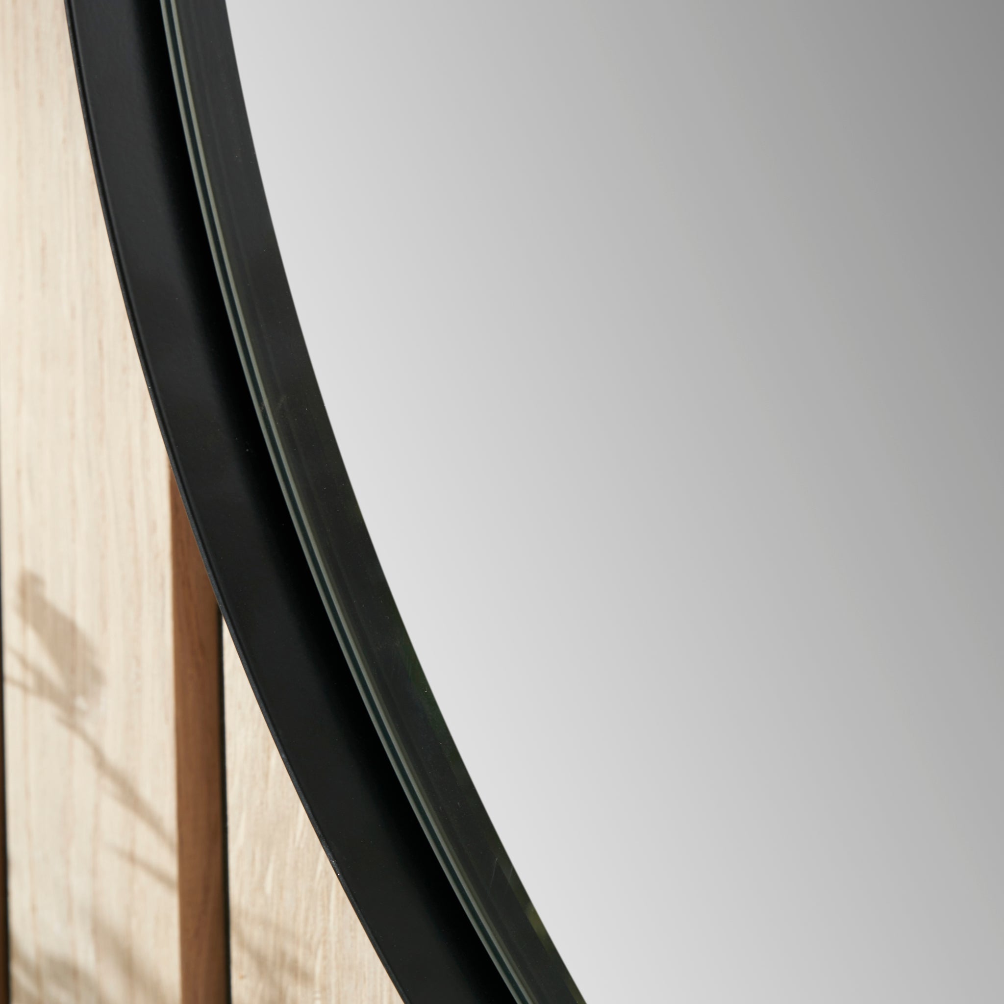A detailed view of the Pacific Lifestyle Black Metal Slim Frame Round Wall Mirror Medium, showcasing its reflection of a light-colored wooden wall.