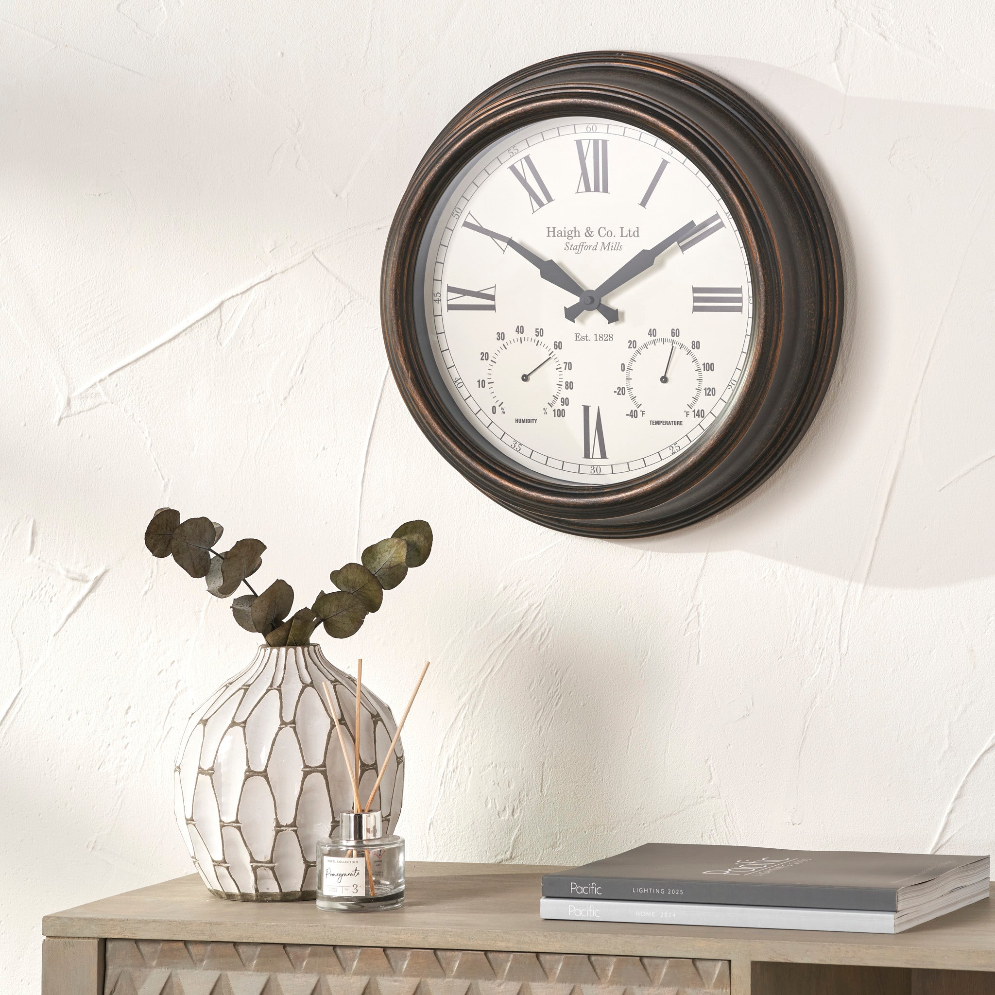 The Pacific Lifestyle - Antique Bronze Effect Indoor Outdoor Wall Clock with Roman numerals and a durable build is shown above a vase with eucalyptus, a reed diffuser, and a closed book.