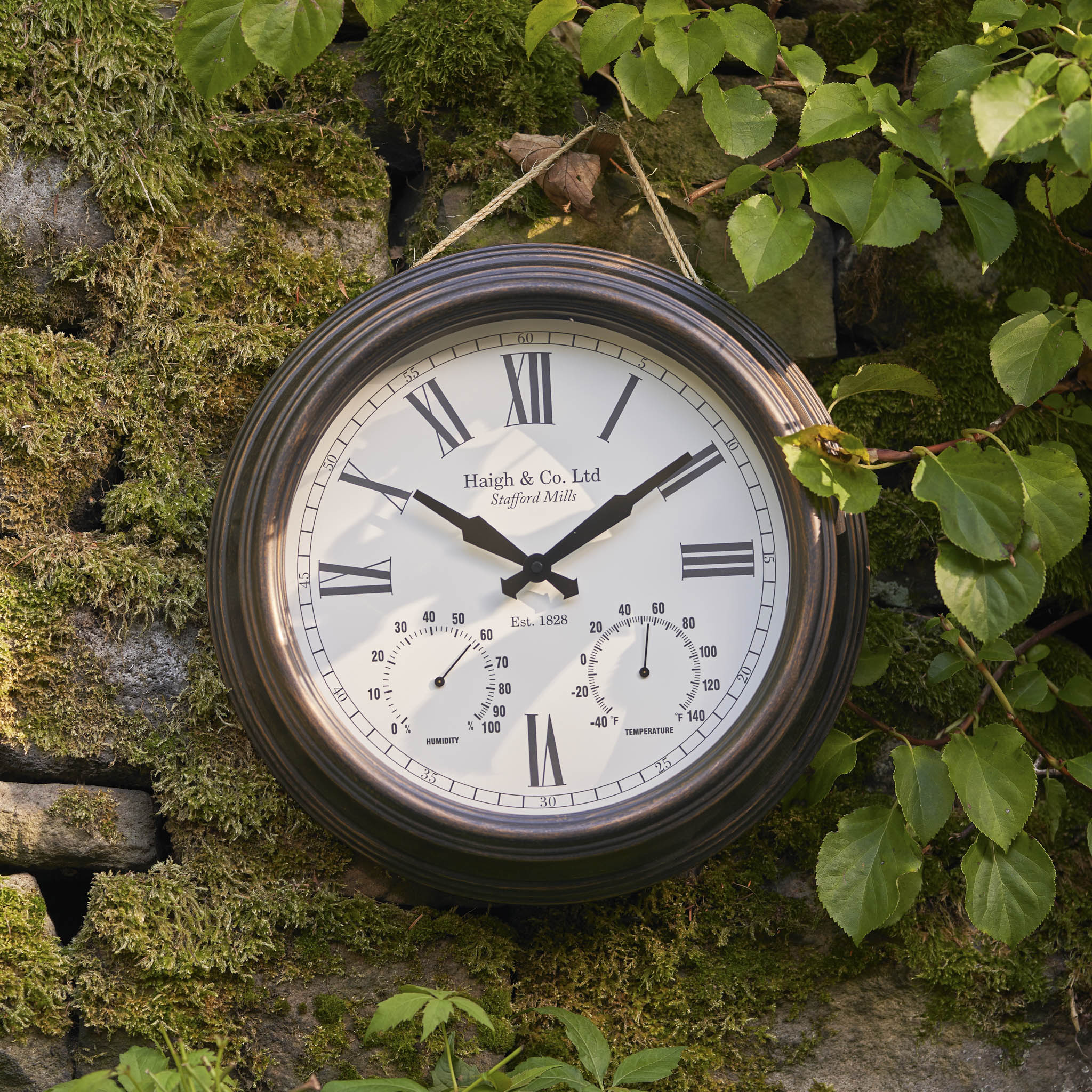 The Pacific Lifestyle - Antique Bronze Effect Indoor Outdoor Wall Clock with Roman numerals hangs on a mossy stone wall amid green leaves, highlighting its durable construction and classic style.