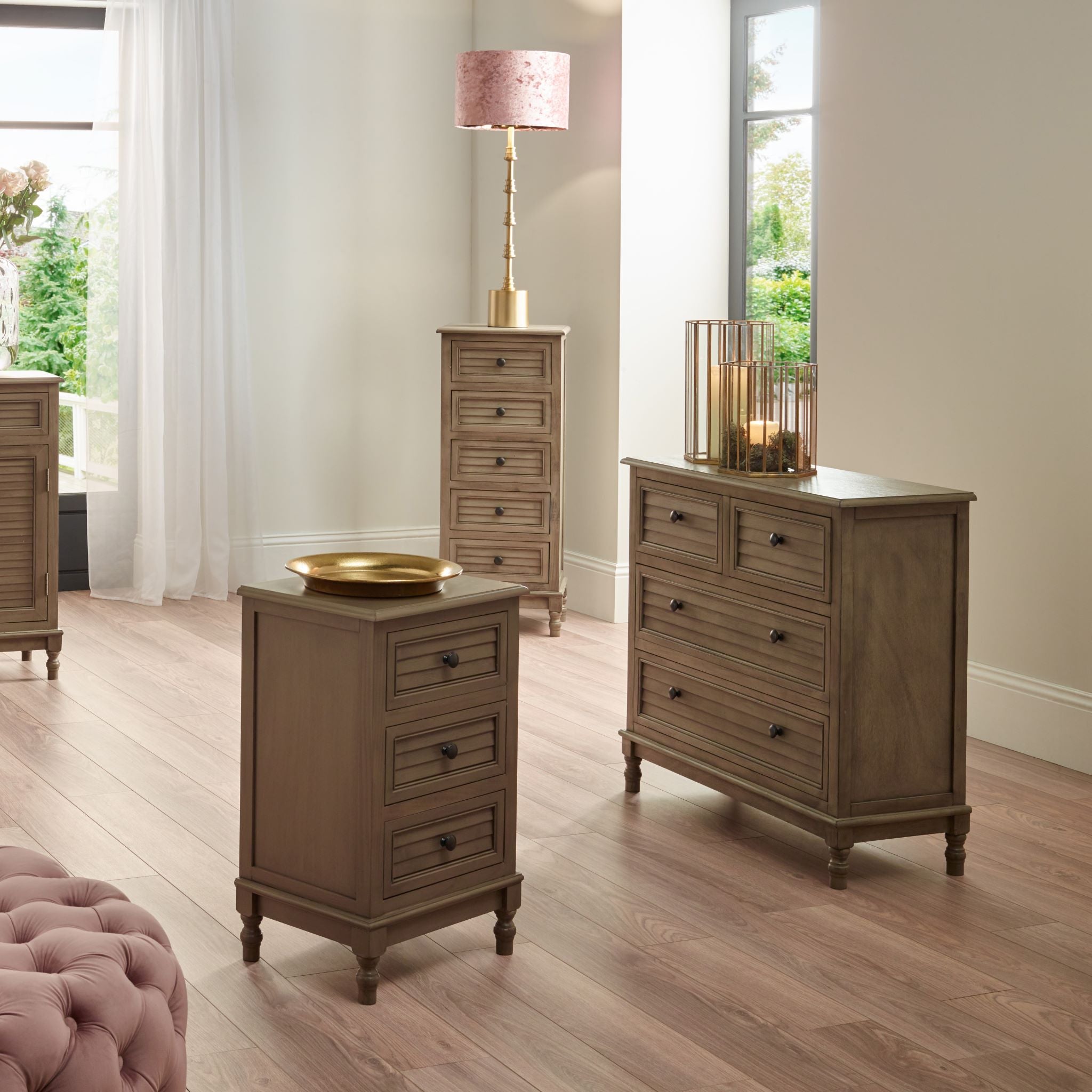 The modern bedroom showcases the Pacific Lifestyle Ashwell Taupe Pine furniture, featuring a 3 Drawer Wood Unit and dressers, paired with a sleek lamp on a light wood floor. Large windows enhance the space's elegance and chic aesthetic.