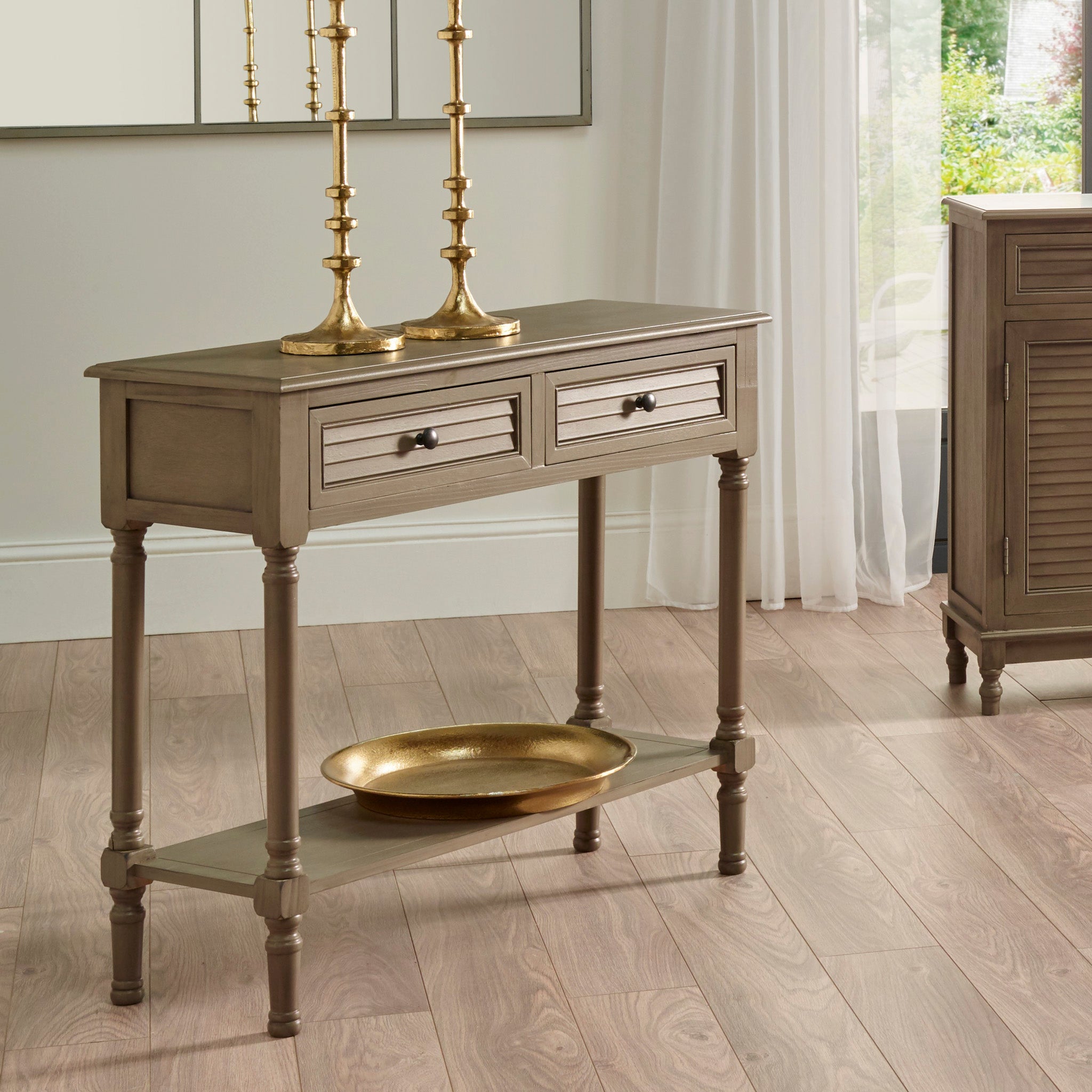 The Pacific Lifestyle Ashwell Taupe Pine Wood Console Table features two drawers, a taupe finish, brass candlesticks, and a decorative gold tray underneath.