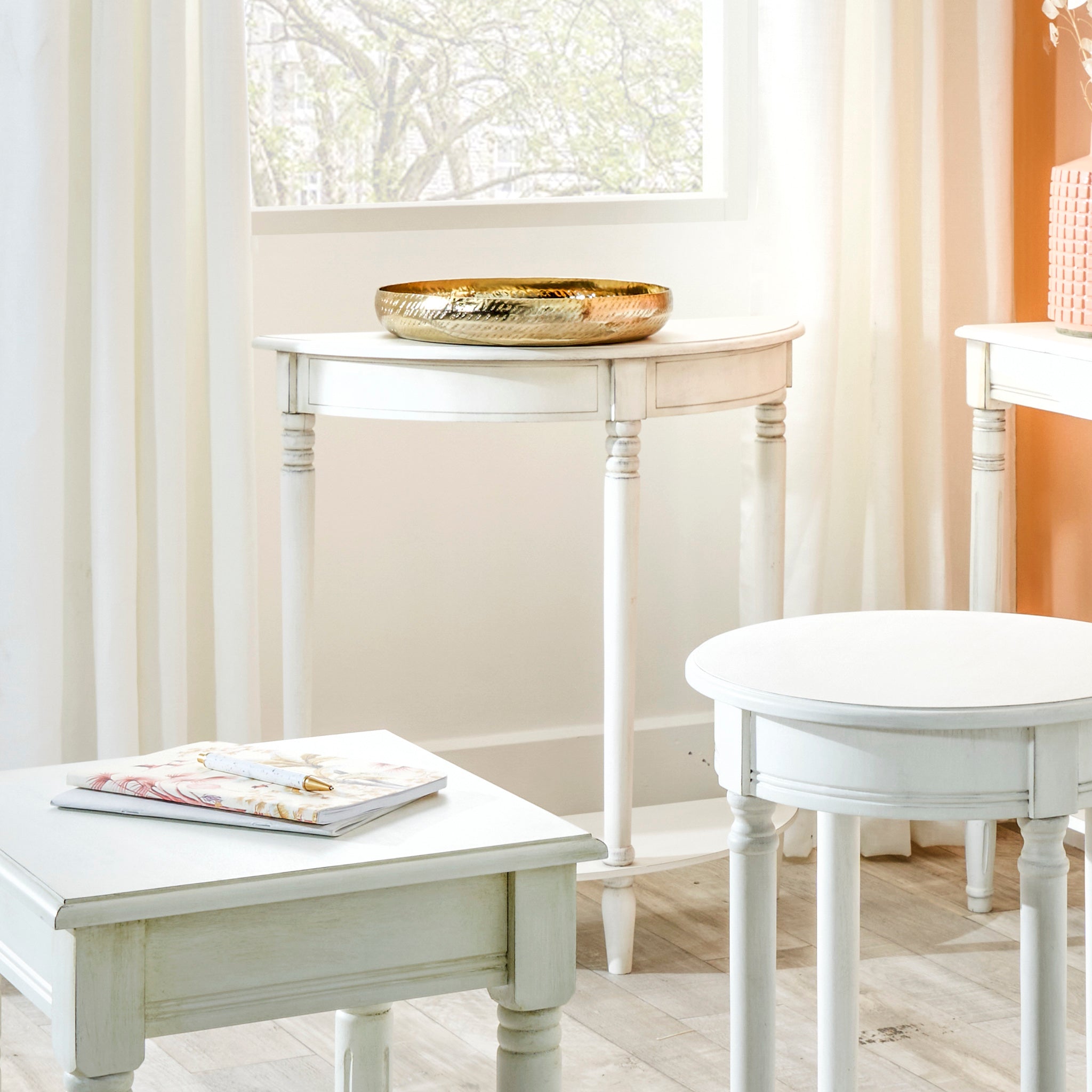 The Pacific Lifestyle Heritage Elizabeth White Pine Wood Half Moon Console Table is styled with a gold bowl and magazine near a window adorned with sheer curtains, offering a tree-filled view.