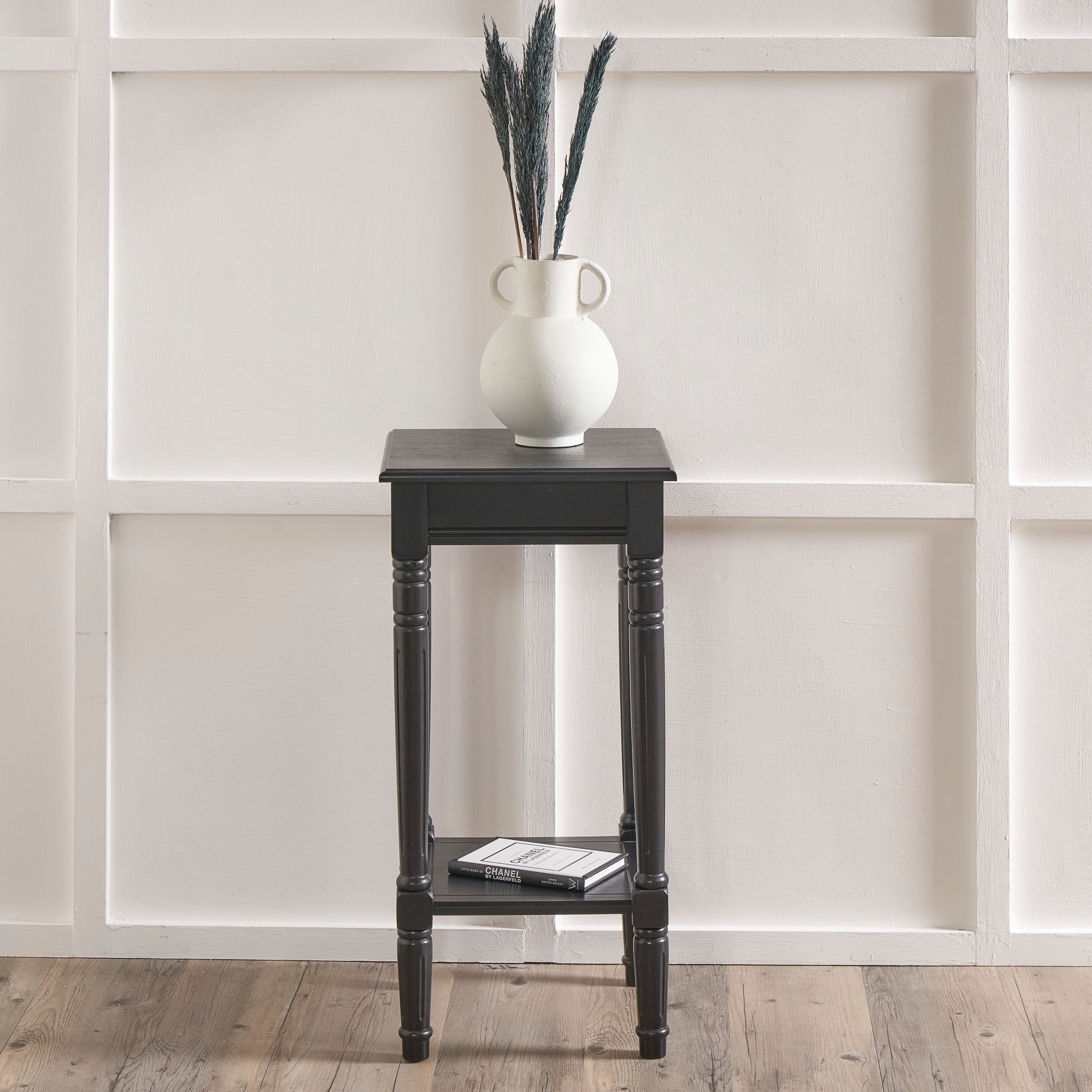 The Pacific Lifestyle - Heritage Black Pine Wood Accent Table showcases a white vase with dried plants against a white paneled wall on a wooden floor.