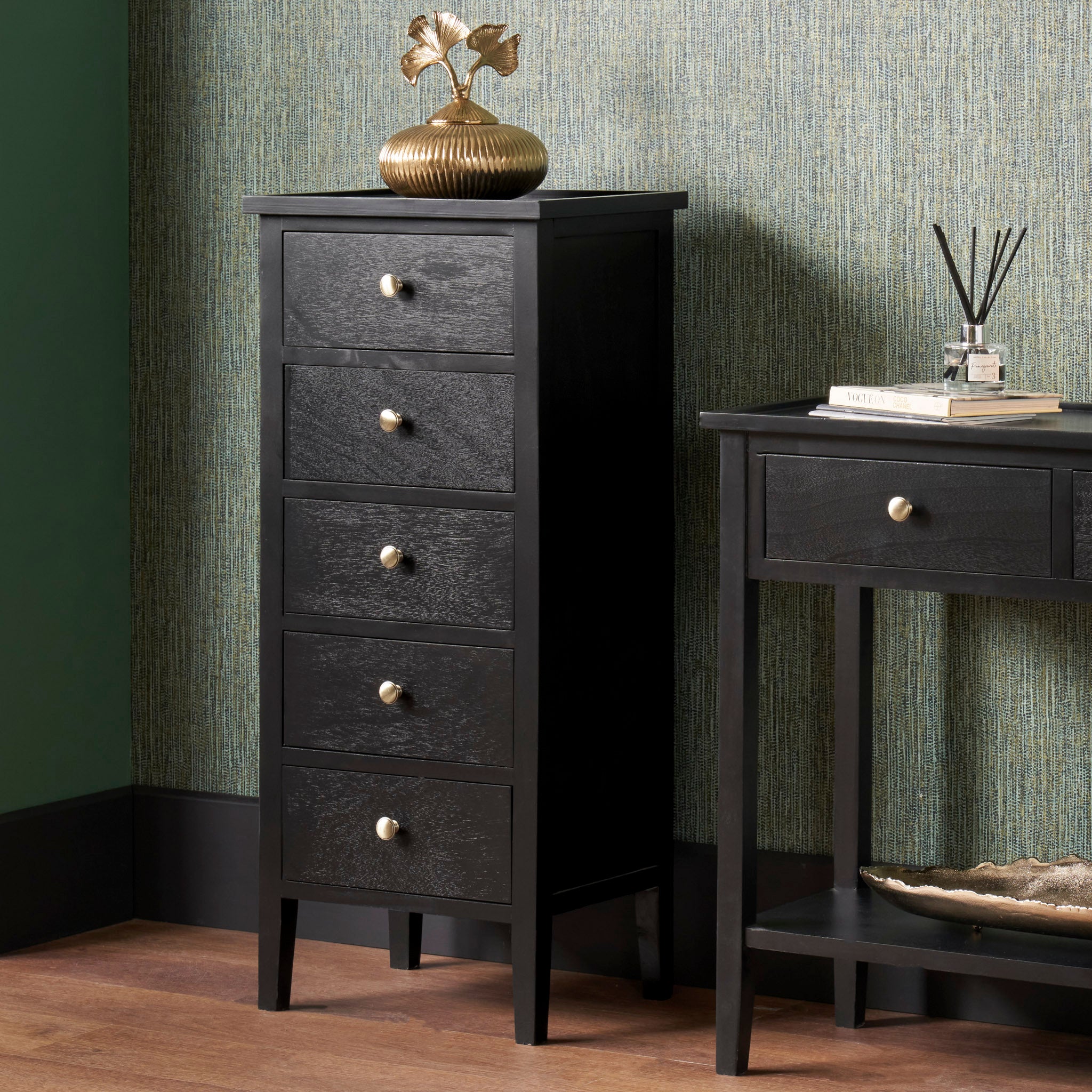 The Pacific Lifestyle Fairford Satin Black Pine Wood 5 Drawer Tall Boy features a gold vase on top, next to a dark console table against green walls, adding a subtle mid-century aesthetic to your space.