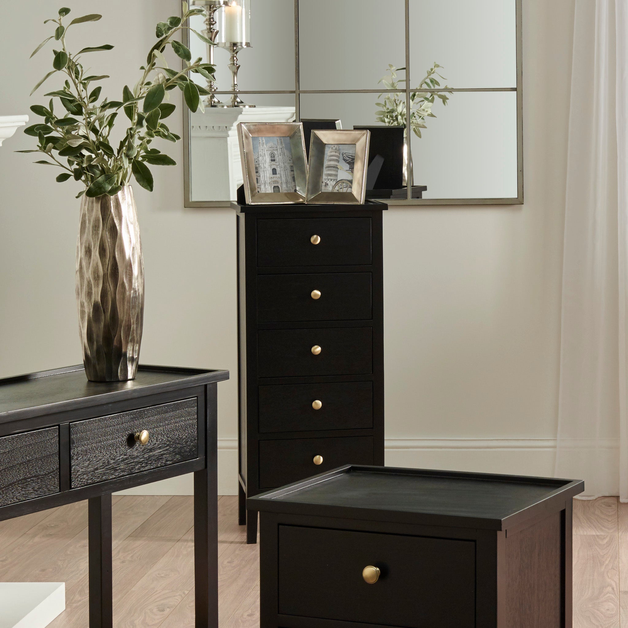 Elegant room featuring the Fairford Satin Black Pine Wood Tall Boy, a textured vase with greenery, framed photos, and a large mirrored wall display for a mid-century look.