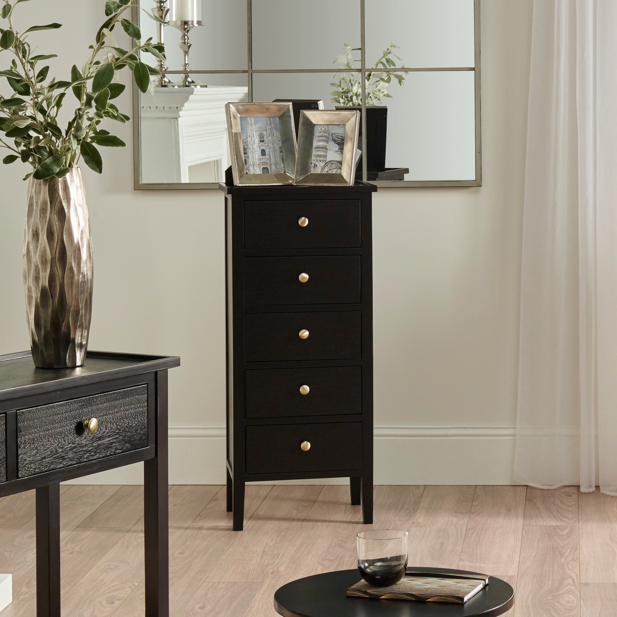 The Pacific Lifestyle Fairford Satin Black Pine Wood 5 Drawer Tall Boy with gold knobs enhances the minimalist room, featuring a vase and framed pictures on top. A mirrored wall complements the mid-century style.