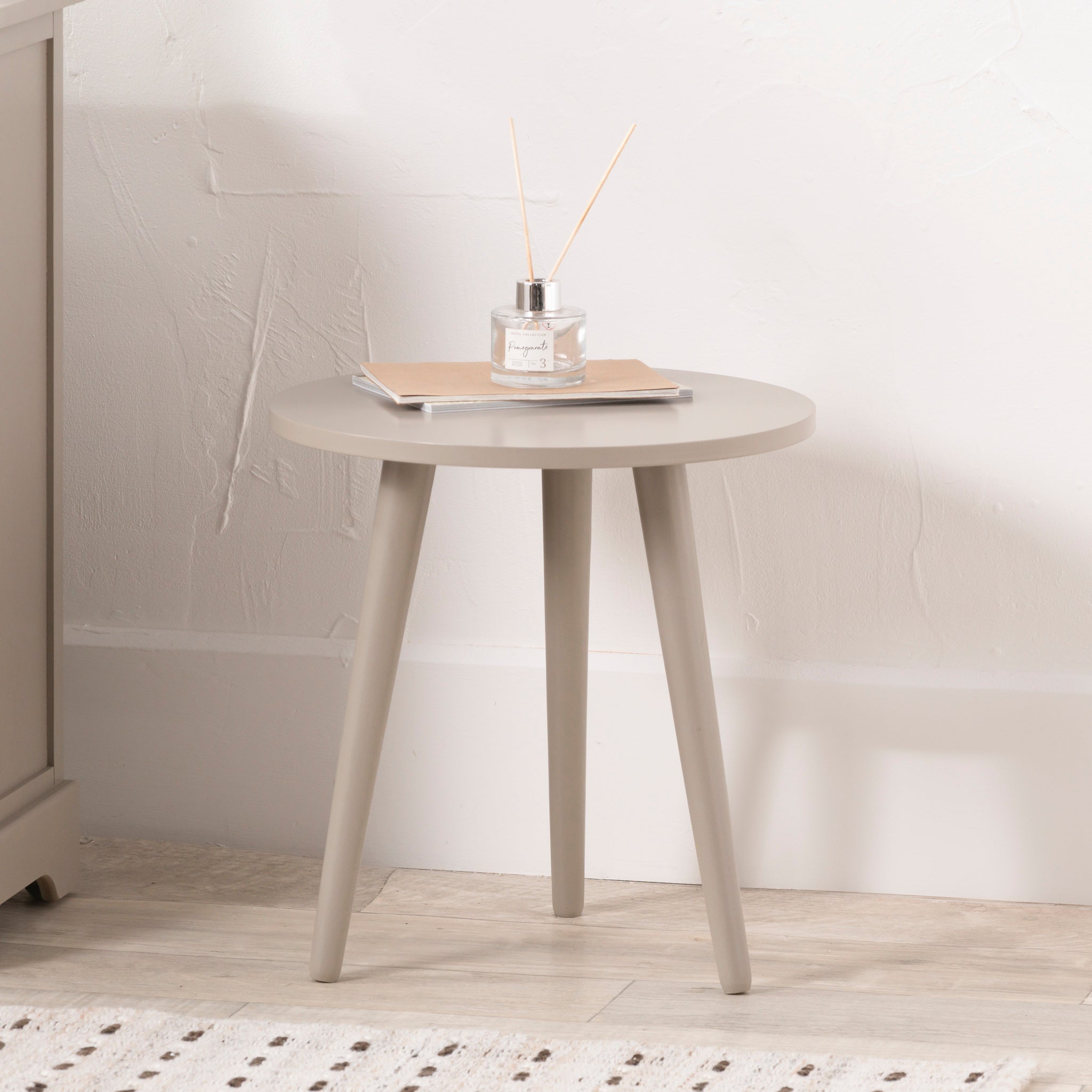 The Pacific Lifestyle Devonshire Putty Pine Wood Side Table features a compact, round beige design with three legs and mid-century style, displayed holding a diffuser and closed book against a white wall.