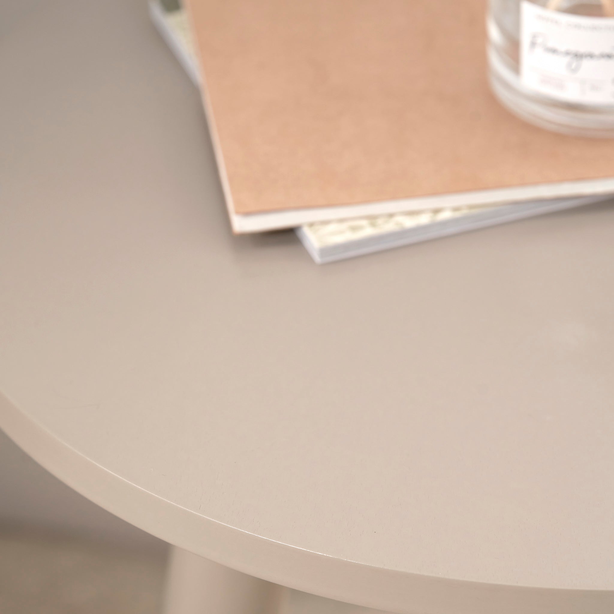 The Pacific Lifestyle Devonshire Putty Pine Wood Side Table, with its round, compact design, is styled with stacked notebooks and a glass jar in the corner for a touch of mid-century charm in any room.