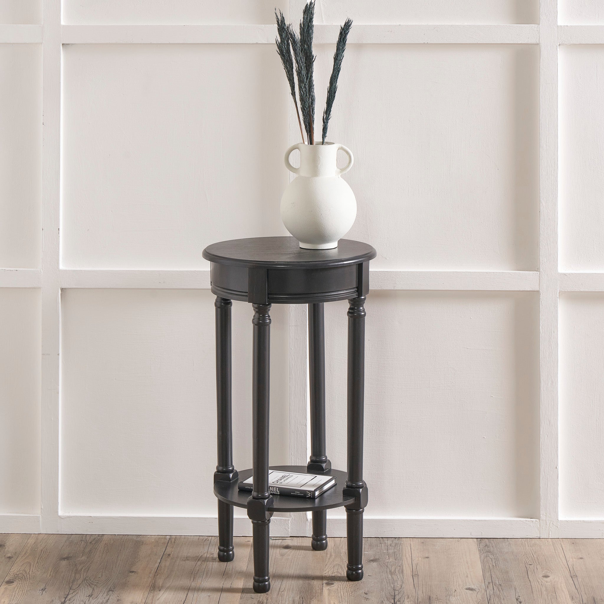 The Heritage Collection showcases the Pacific Lifestyle - Heritage Black Pine Wood Round Accent Table paired with a white vase, enhancing the elegance of a paneled white wall and wooden floor.