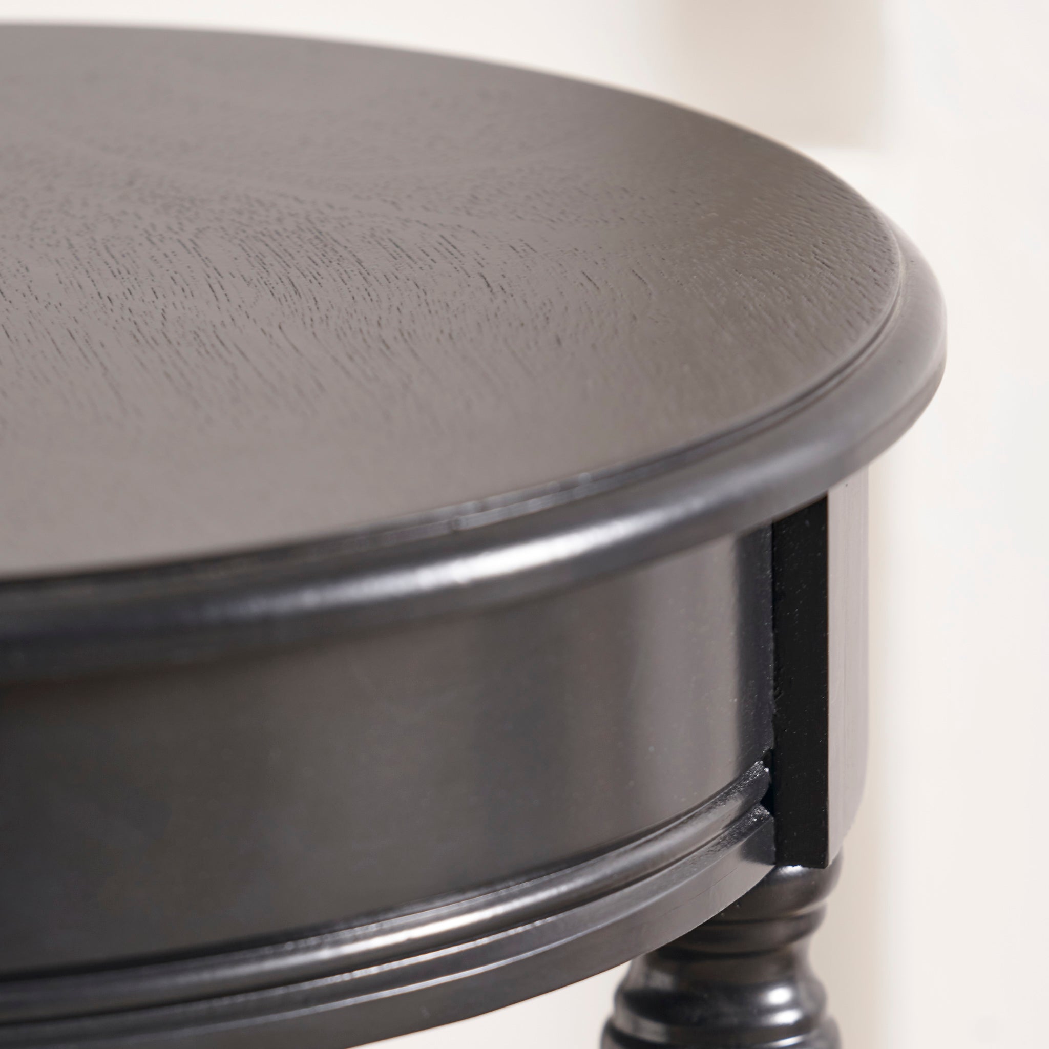 This close-up features the Pacific Lifestyle - Heritage Black Pine Wood Round Accent Table, highlighting its detailed edge. This elegant piece from the Heritage Collection embodies the timeless allure of black lacquered furniture.