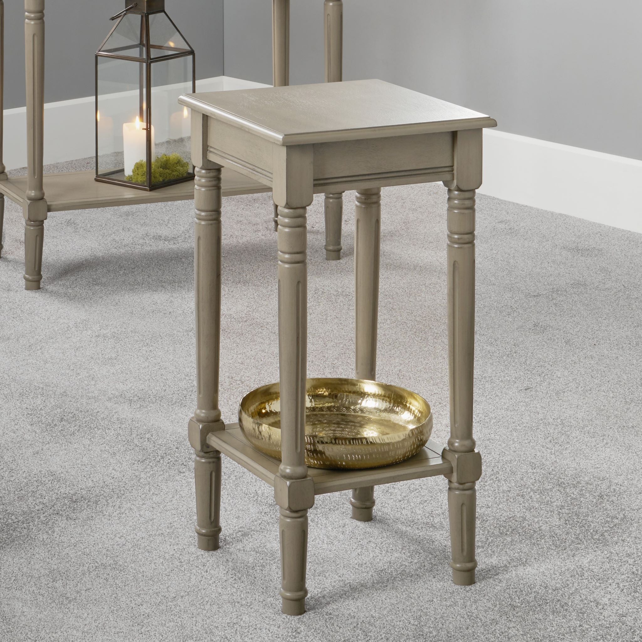 The Pacific Lifestyle - Heritage Taupe Pine Wood Square Accent Table features decorative legs and a lower shelf with a gold bowl, while a lantern adds charm in the background.