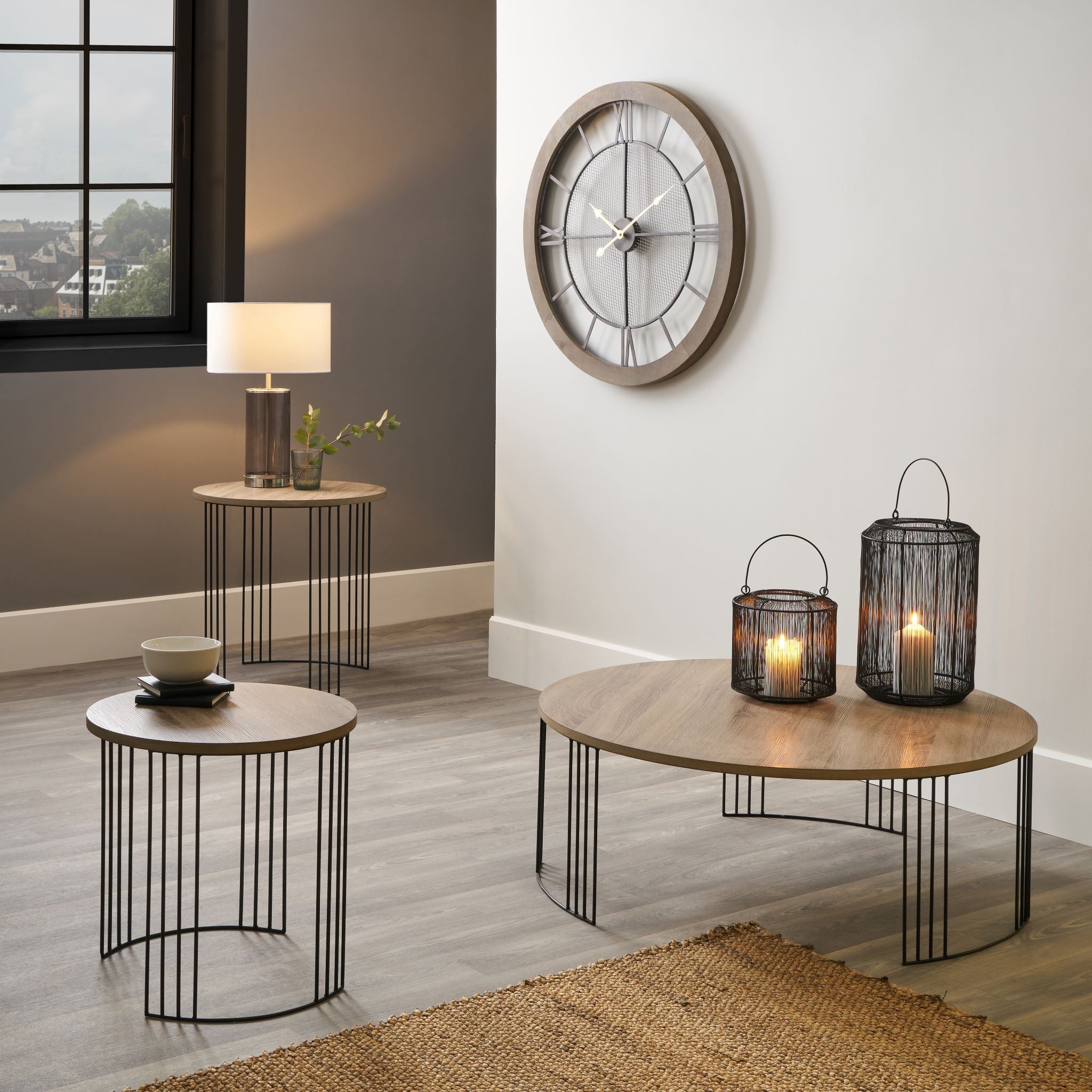 A modern living room showcasing the Pacific Lifestyle Atri dark wood and black metal round coffee table, candles, a lamp, a stylish round wall clock, and a captivating window view.