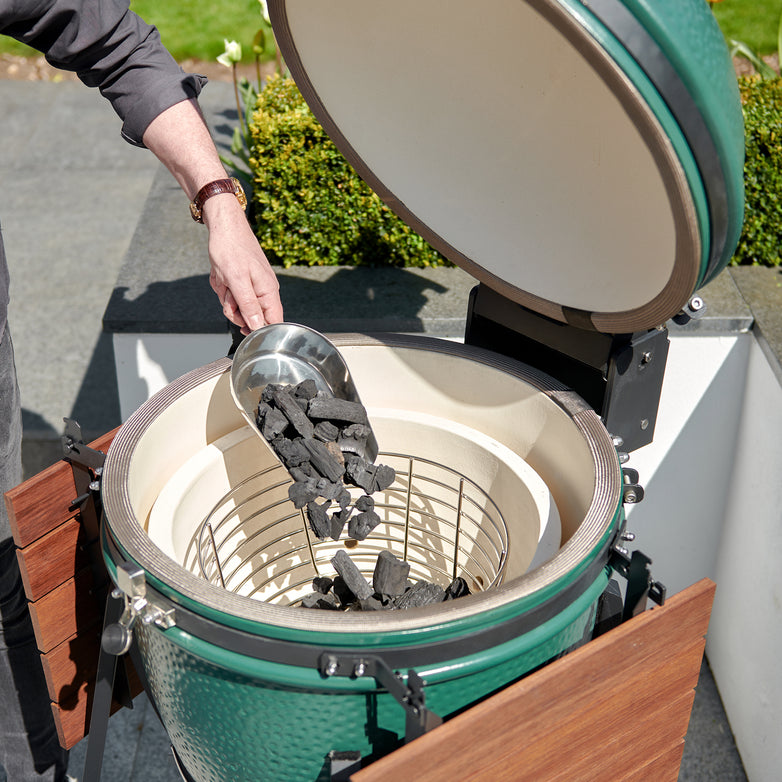 Someone is pouring charcoal into a green ceramic grill in an outdoor setting, with the Kamado Ash Shovel conveniently placed nearby for efficient grill cleaning later.