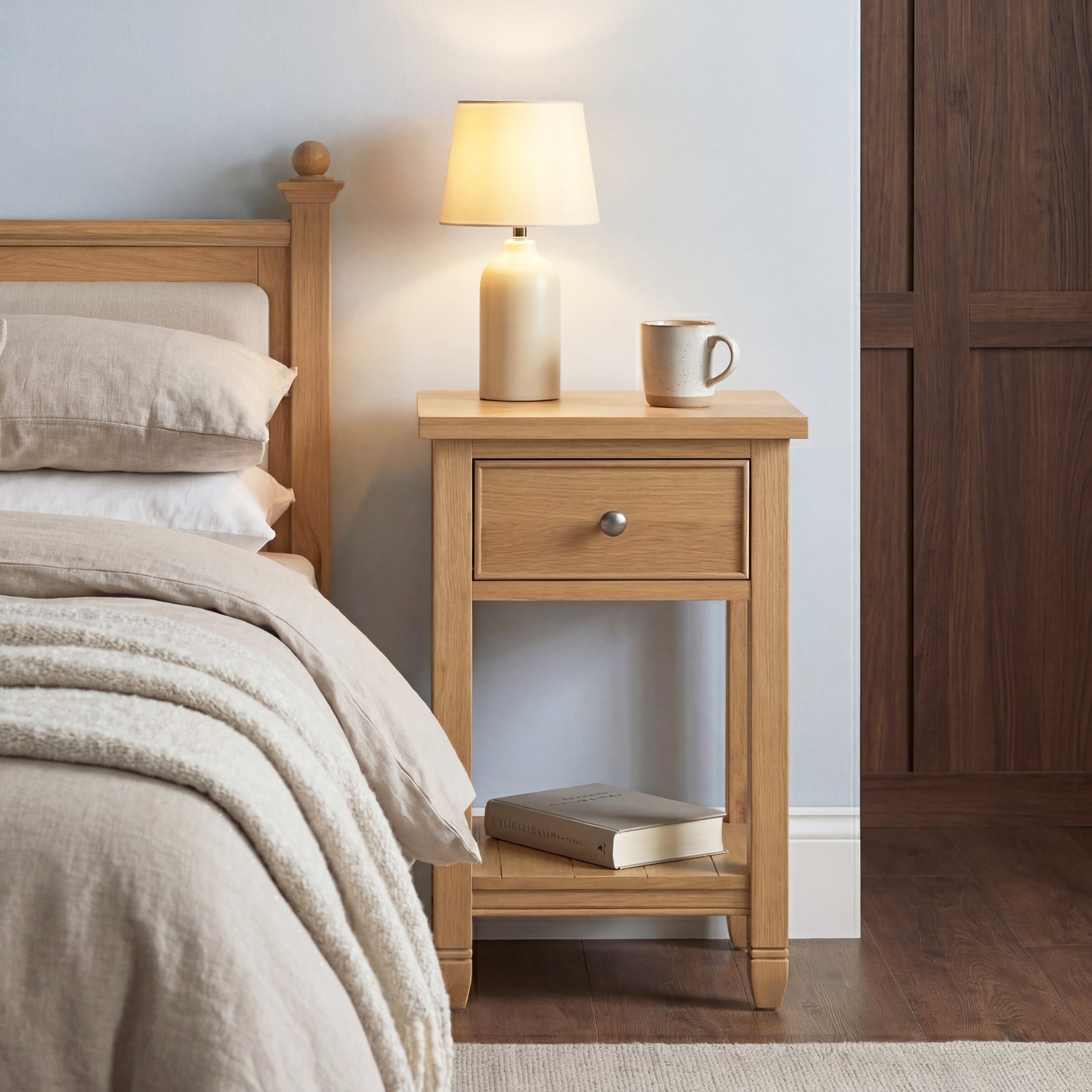 An Amberley Bedside Table in Light Oak sits next to a neatly made bed with beige bedding, topped with a lamp, mug, and books.