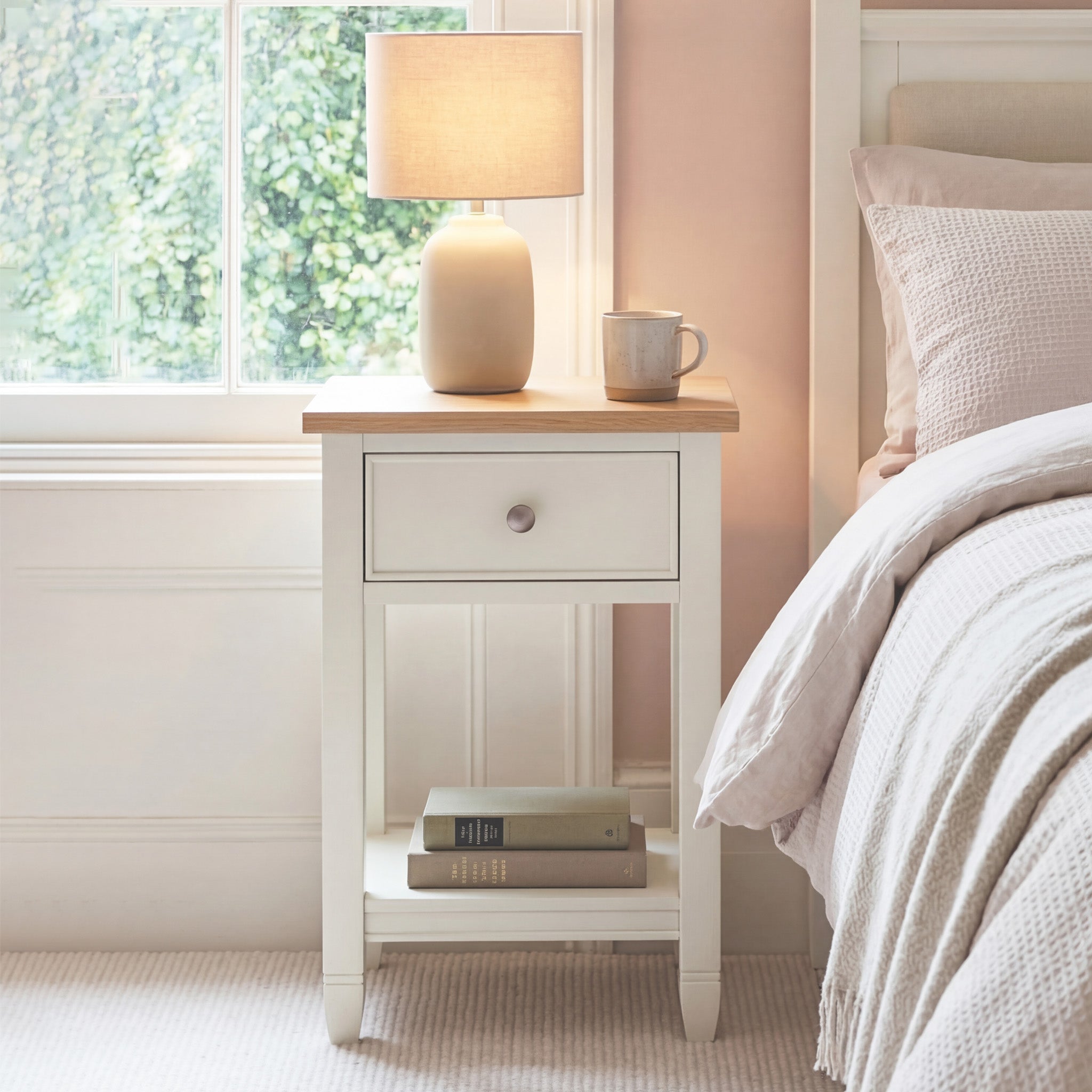 An Amberley Bedside Table in Warm White holds a lamp, mug, and books beside a neatly made bed near a sunny window.
