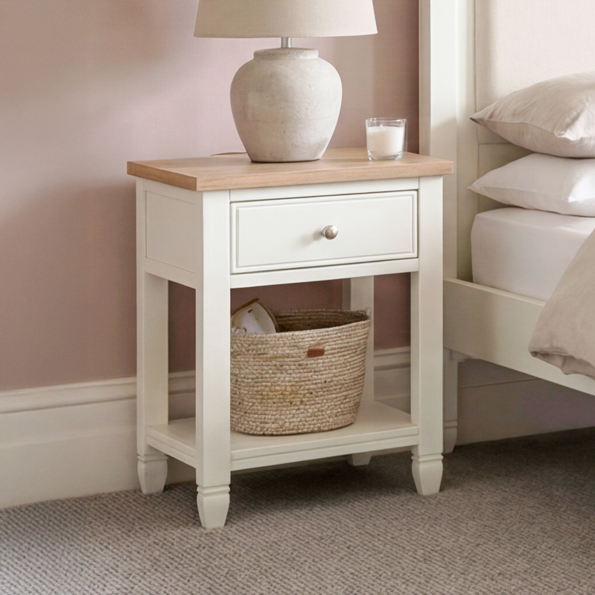 The Amberley Bedside Table in Warm White features a drawer and shelf basket, shown with a lamp and candle beside a bed in a softly lit room—ideal as a compact bedside table for adding modern country charm.
