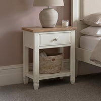 The Amberley Bedside Table in Warm White, with a lamp, candle, and basket on its shelf, stands beside a bed in a softly lit room.
