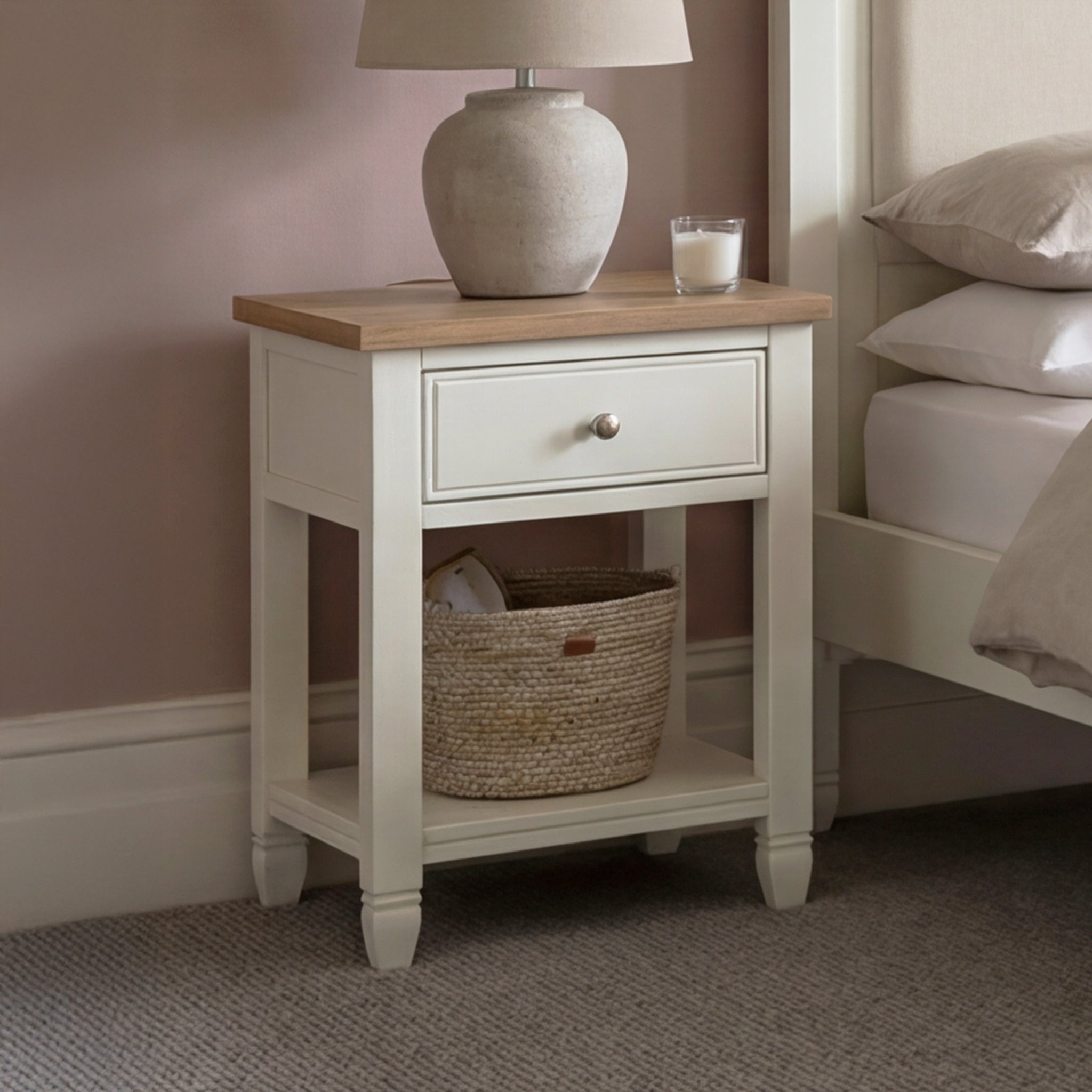 The Amberley Bedside Table in Warm White, with a lamp, candle, and basket on its shelf, stands beside a bed in a softly lit room.