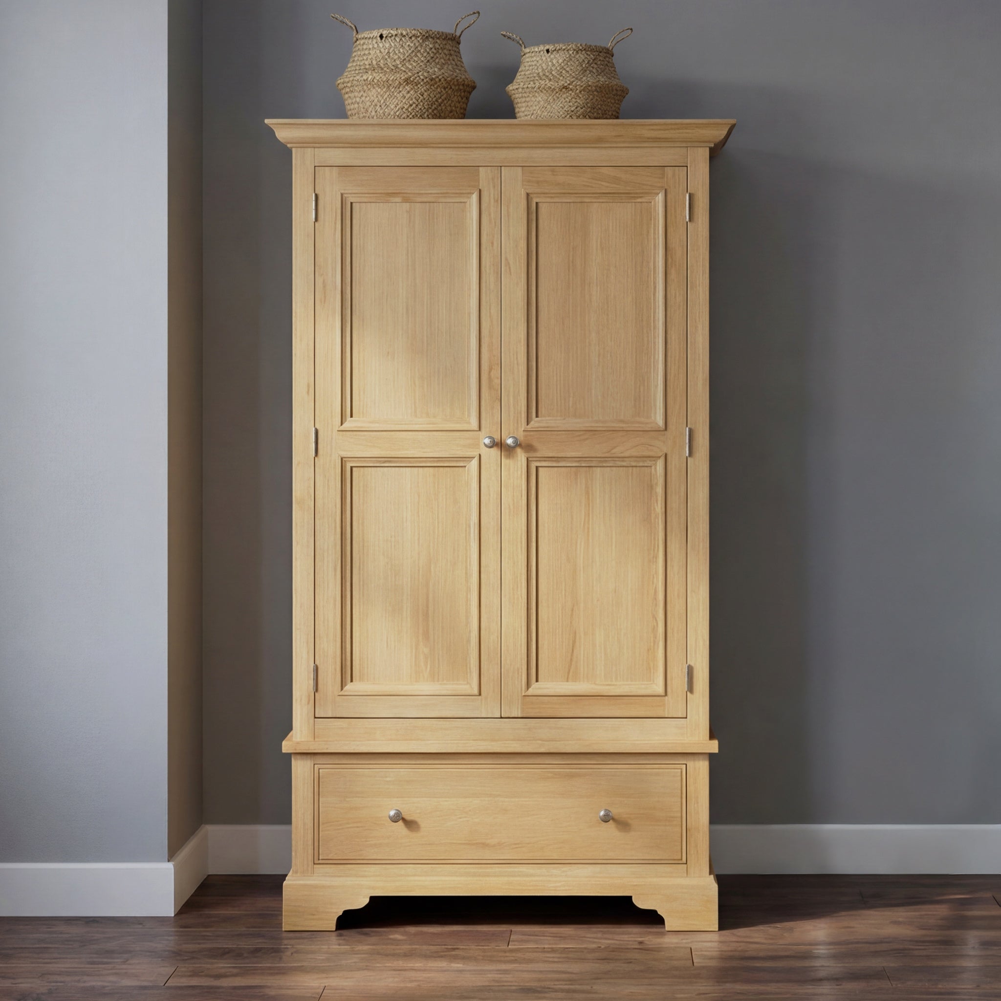 The Amberley Double Wardrobe in Light Oak features a lower drawer and two woven baskets on top, shown against a gray wall and wooden floor.