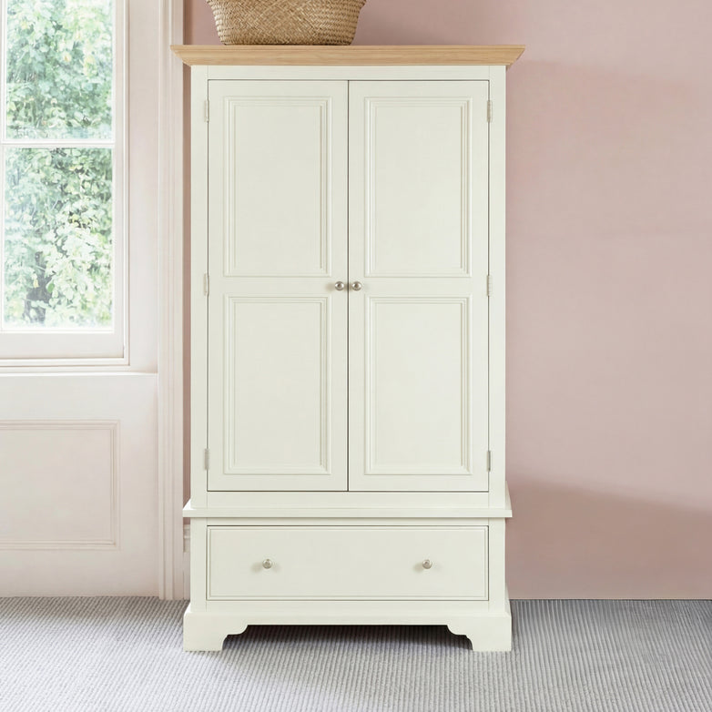The Amberley Double Wardrobe in Warm White features a lower drawer and top basket, set by a pale pink wall near a window, offering elegant bedroom storage.