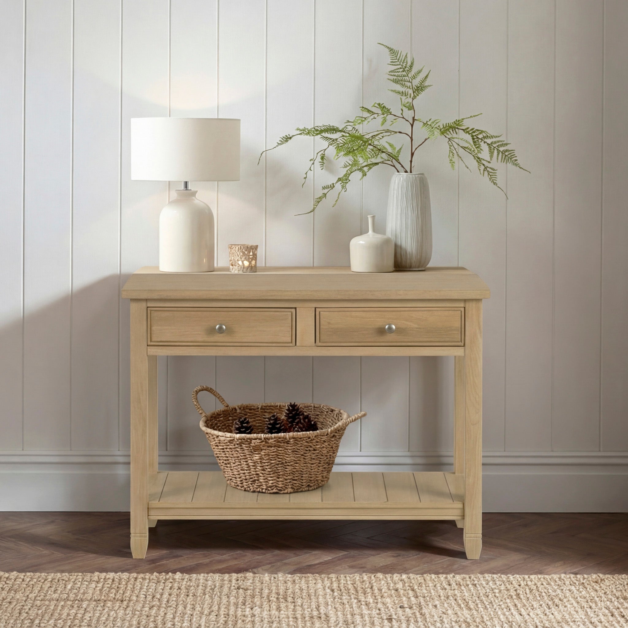 The Amberley 2 Drawers Console Table in Light Oak, styled with a lamp, vases, and basket against a white paneled wall, makes a perfect and stylish hallway addition.