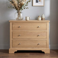 The Amberley 3 Chest of Drawers in Light Oak features three spacious drawers and looks stylish with vases and dried flowers on top—ideal bedroom storage furniture against a light gray wall.