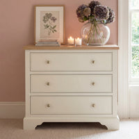 The Amberley 3 Chest of Drawers in Warm White offers compact bedroom storage and is styled with a vase of flowers, candles, books, and a framed botanical print on top.