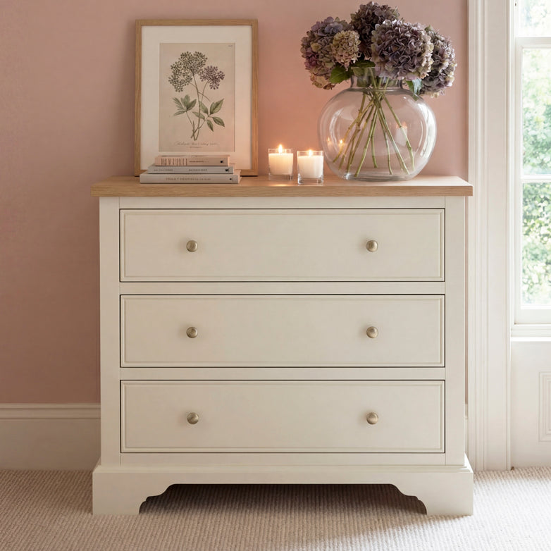The Amberley 3 Chest of Drawers in Warm White offers compact bedroom storage and is styled with a vase of flowers, candles, books, and a framed botanical print on top.