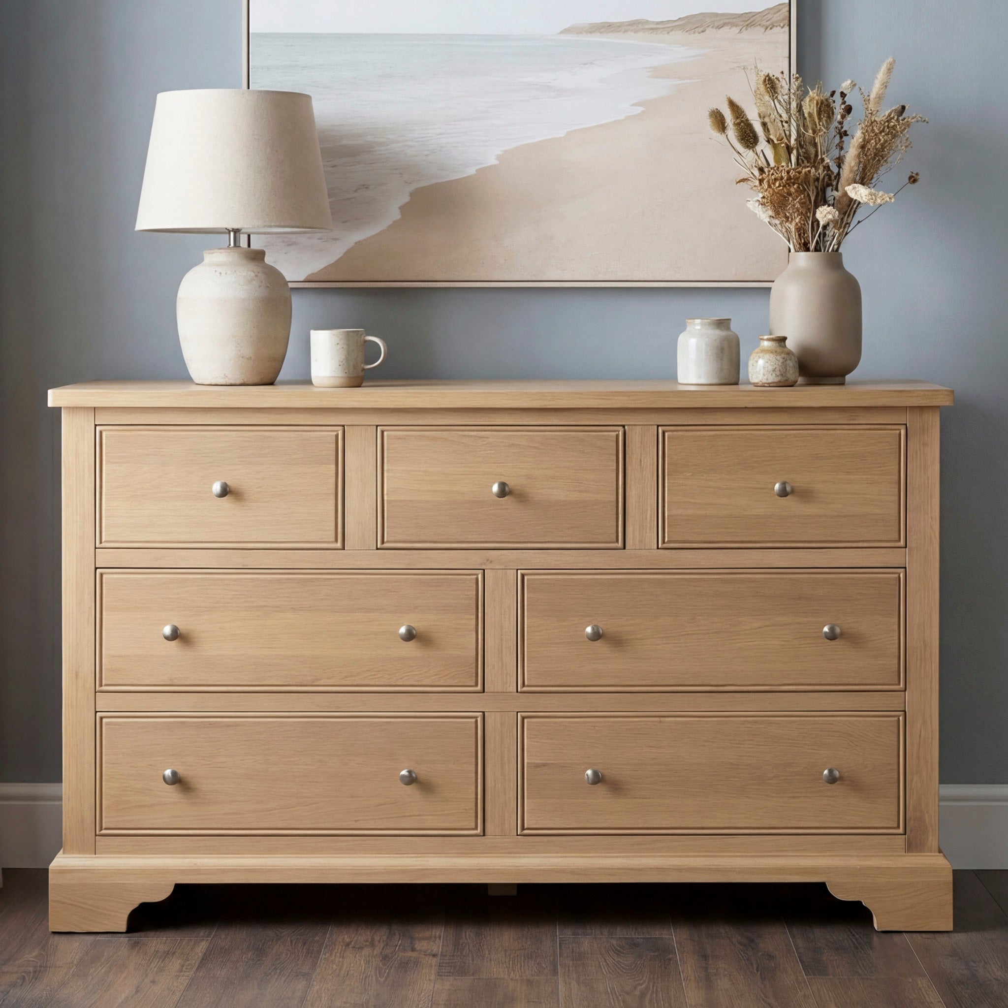 The Amberley 3 Over 4 Chest of Drawers in Light Oak features seven drawers, ideal for bedroom storage, and is shown styled with a lamp, vases, a mug, and beach artwork above.
