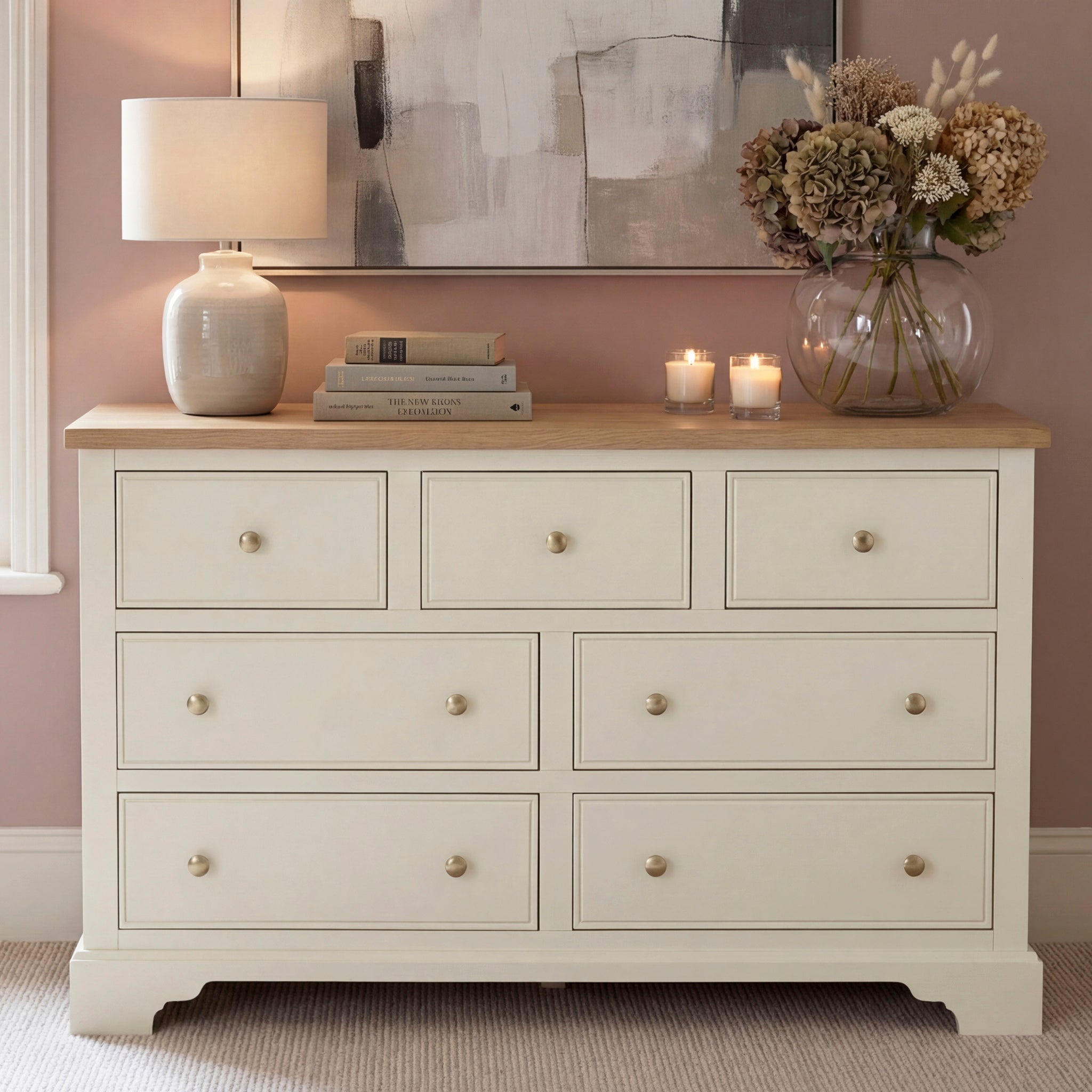 The Amberley 3 Over 4 Chest of Drawers in Warm White features an oak top and seven drawers, styled with a lamp, books, candles, and a vase of flowers set against a pink wall.