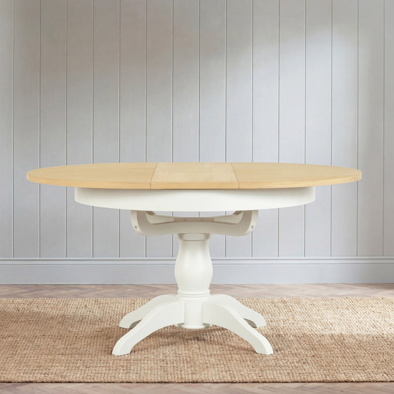 The Amberley 4-6 Seater Round Extending Dining Table in Warm White, featuring a light wood top and white pedestal base, is placed on a woven rug in a bright room.