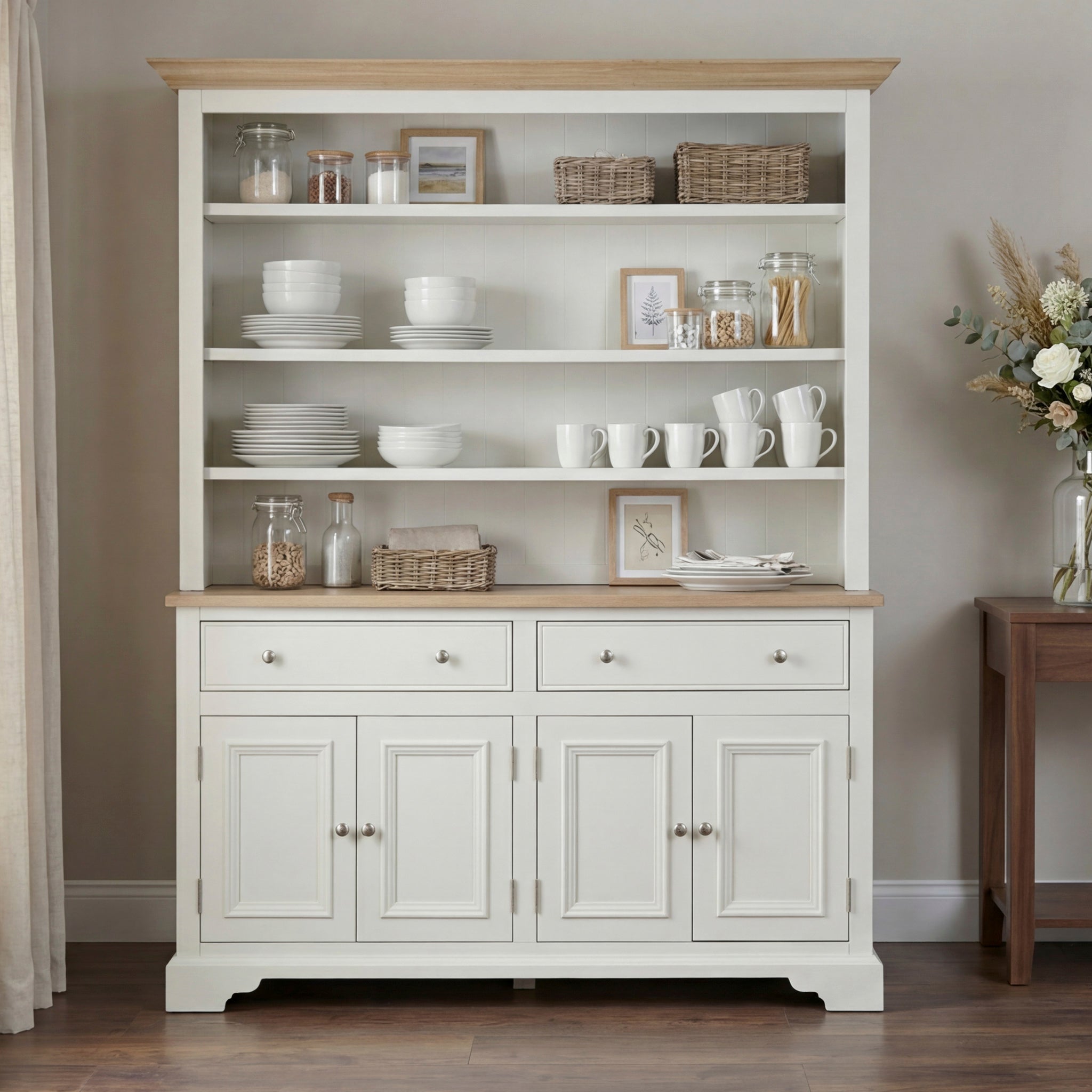 The Amberley 5ft Grand Open Dresser in Warm White showcases dishes, mugs, baskets, and decor on its shelves; flowers on a nearby table highlight its classic English craftsmanship.