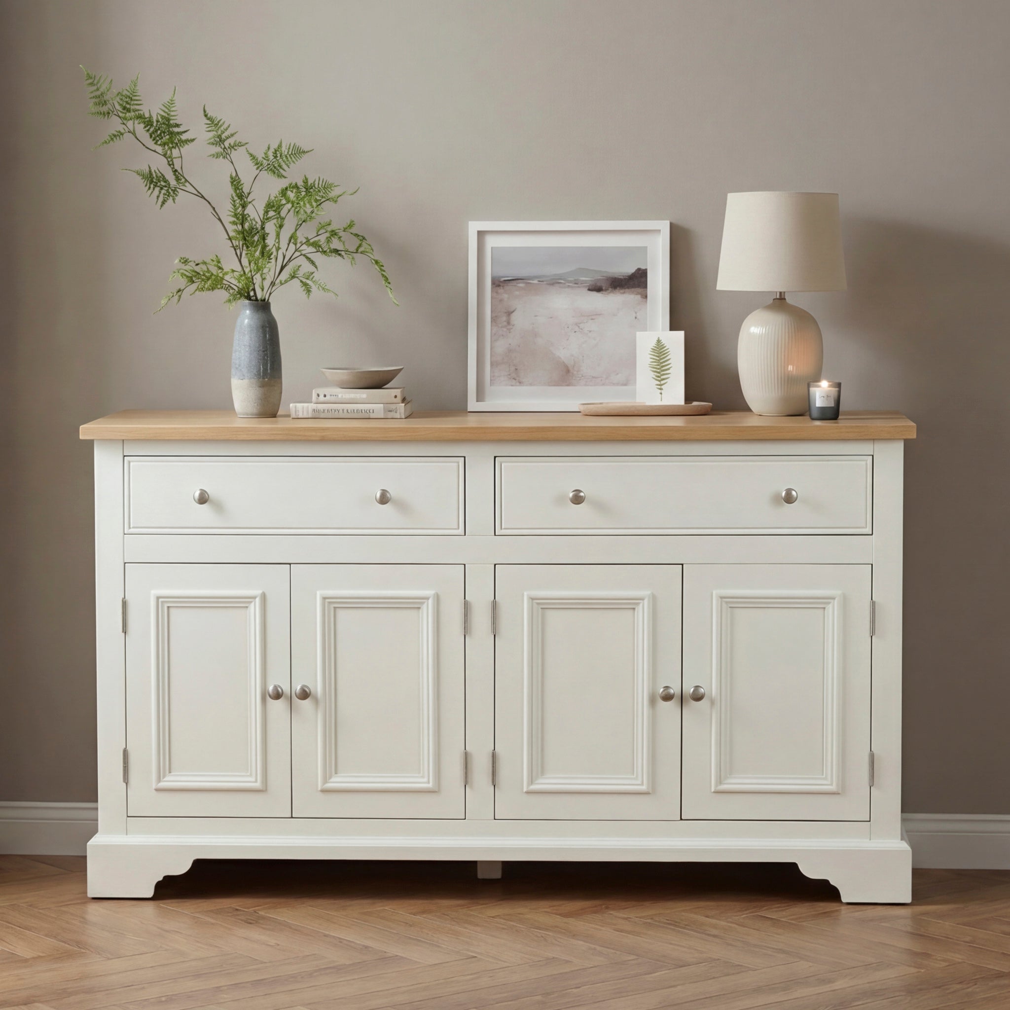 The Amberley Large 4 Door 2 Drawers Sideboard in Warm White, topped with decor items like a lamp, vase, art, and a plant.