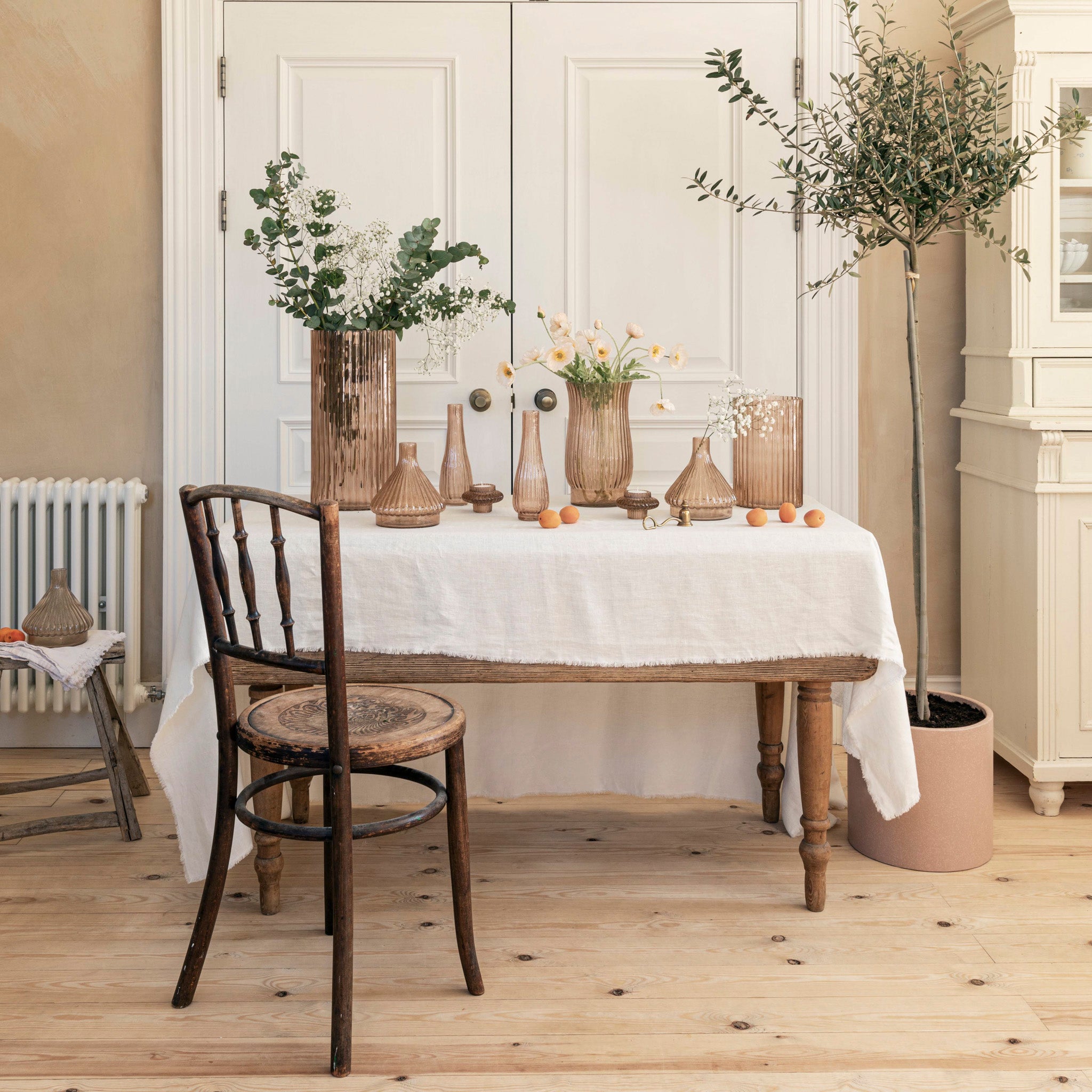 Rustic room featuring a sustainable wooden table, linen tablecloth, Ivyline – Daphne Large Ribbed Vase in Apricot, plants, and a wooden chair on a light wood floor.