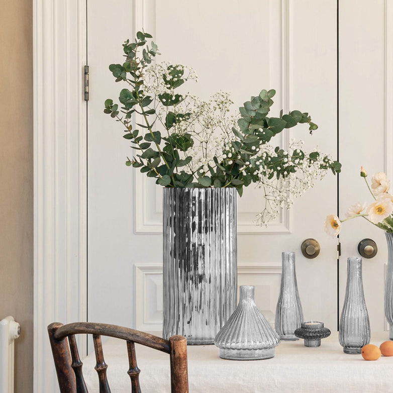 The Ivyline - Daphne Large Ribbed Vase in Clear, topped with greenery and flowers, adds elegance to the table décor beside a white paneled door and a wooden chair.